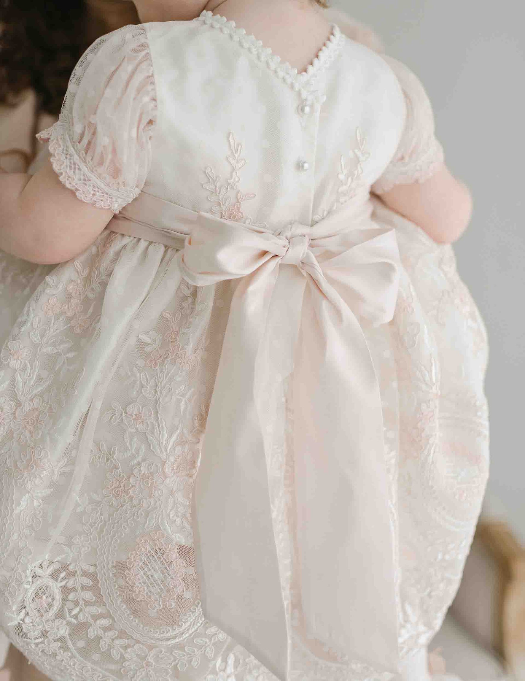 A baby is dressed in the Elizabeth Christening Dress, which features intricate lace detailing and a large satin bow at the back. This handmade white dress showcases short puff sleeves and decorative pearl buttons along the neckline. The baby’s back is facing the camera.