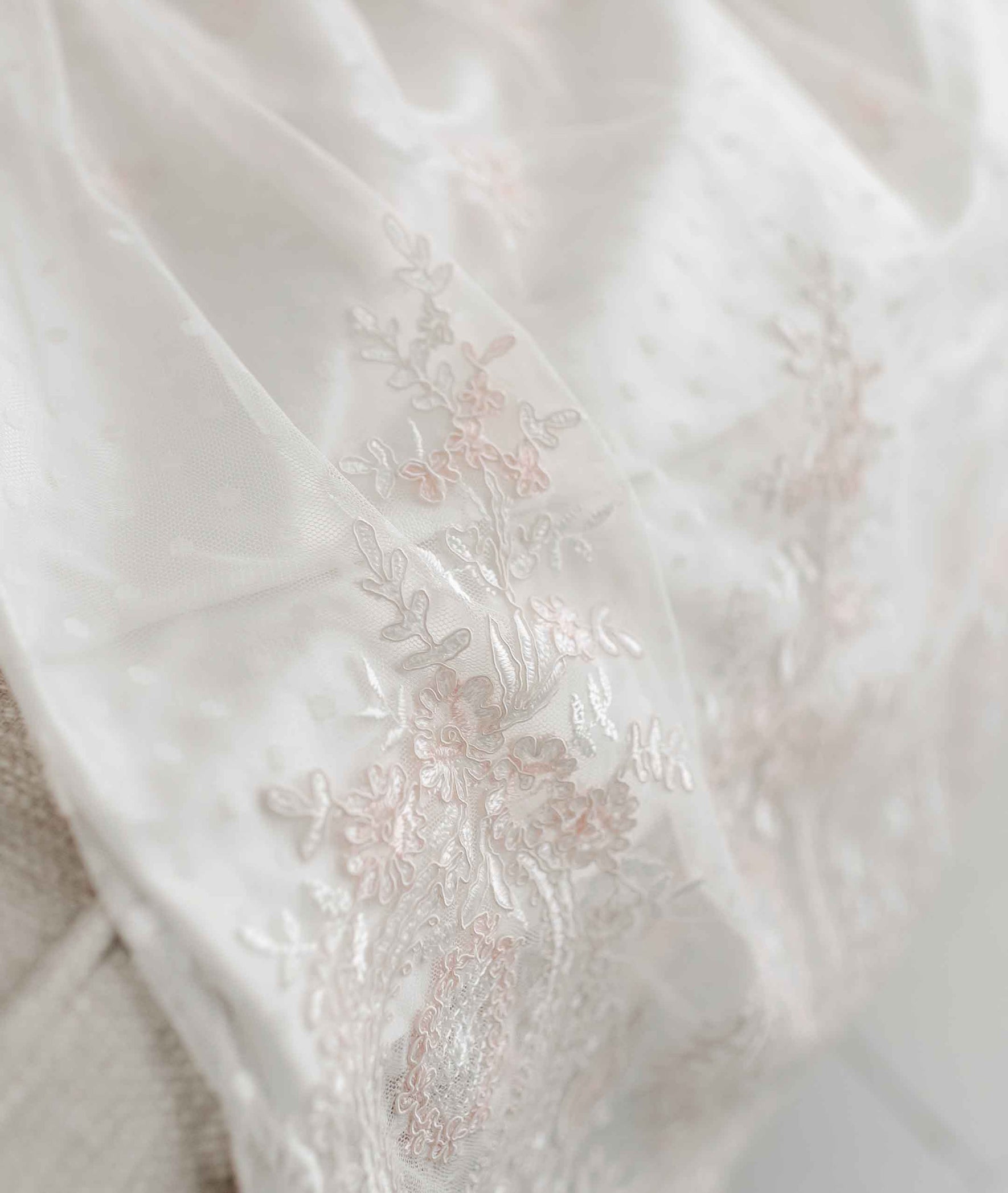 A close-up of the handmade Elizabeth Christening Gown & Bonnet features white silk Dupioni fabric adorned with delicate floral embroidery in light pink and white threads. The fabric has a subtle sheen and appears to be draped or laid flat.
