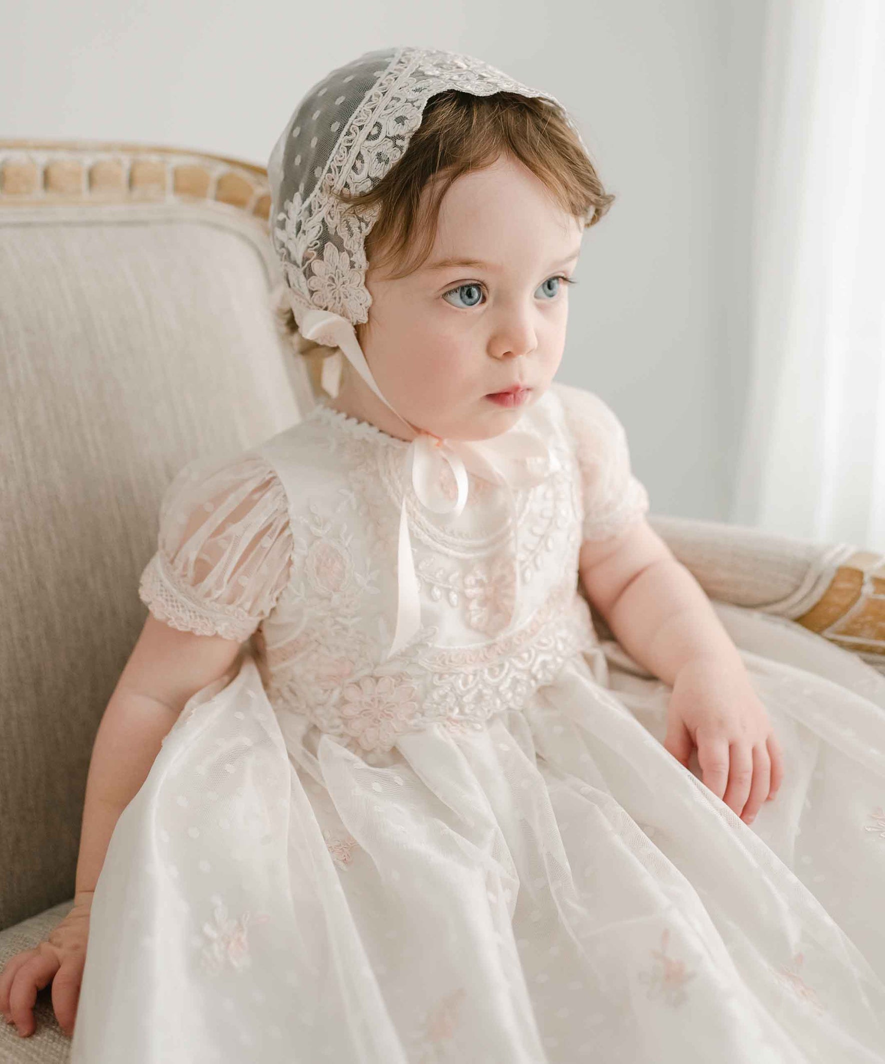 A young child wearing the Elizabeth Christening Gown & Bonnet sits in a beige upholstered chair. The gown, made from exquisite silk Dupioni, enhances the gentle, serene atmosphere. The child looks to the side with a calm expression, bathed in soft light.