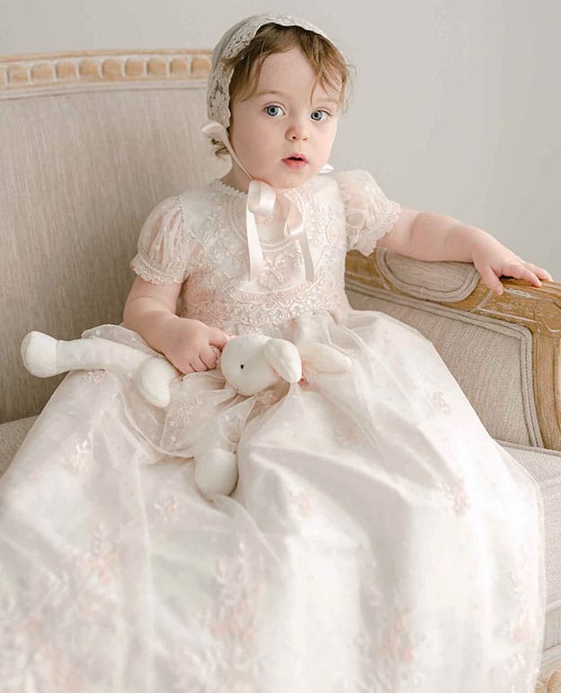 A toddler wearing the Elizabeth Christening Gown and Bonnet sits on an upholstered chair, holding a stuffed white bunny and looking toward the camera.