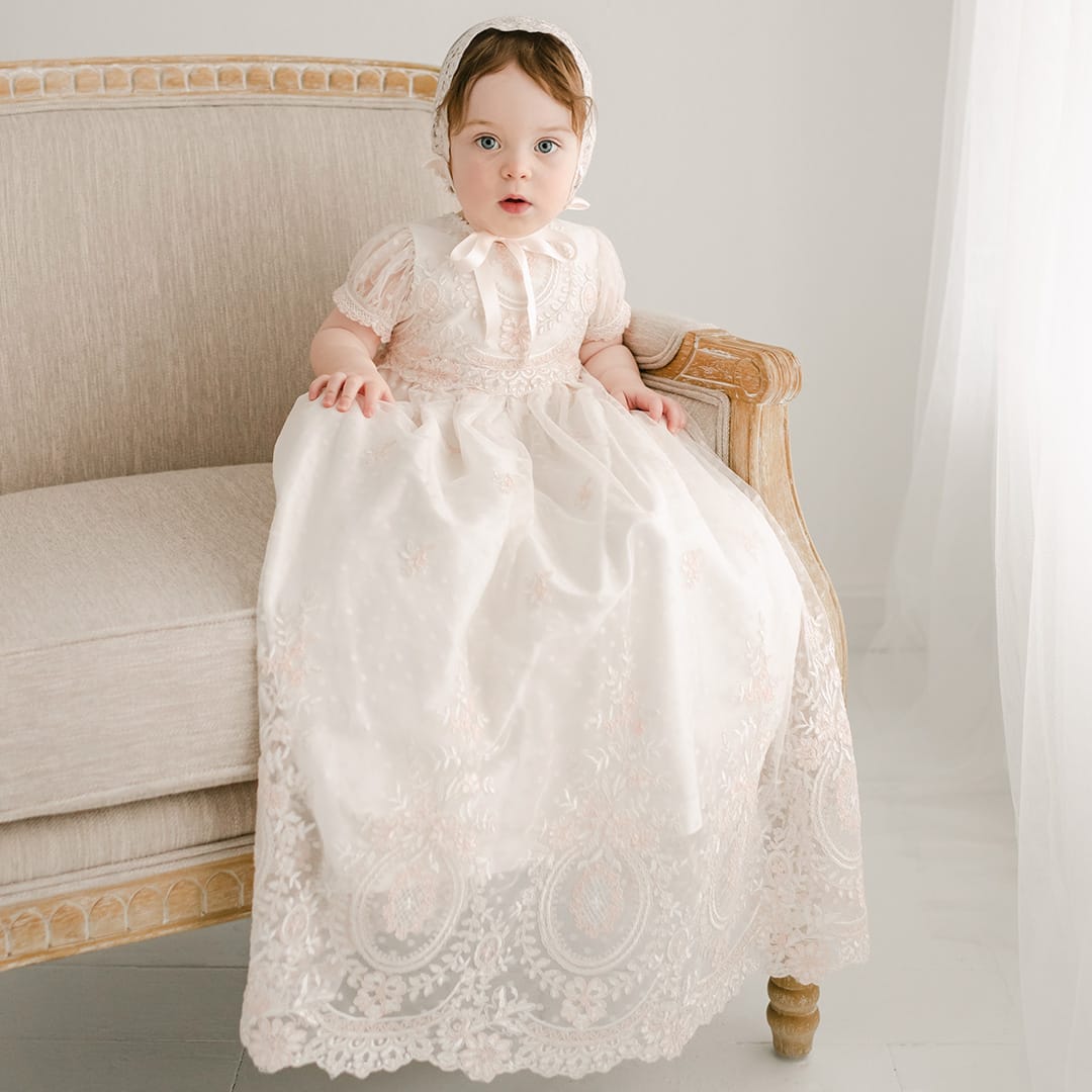 A baby in the Elizabeth Christening Gown & Bonnet, crafted from silk Dupioni with lace detailing, sits on a beige armchair. The baby's curious expression meets the camera's gaze. The background is softly lit and neutral-toned, enhancing the tranquil scene.