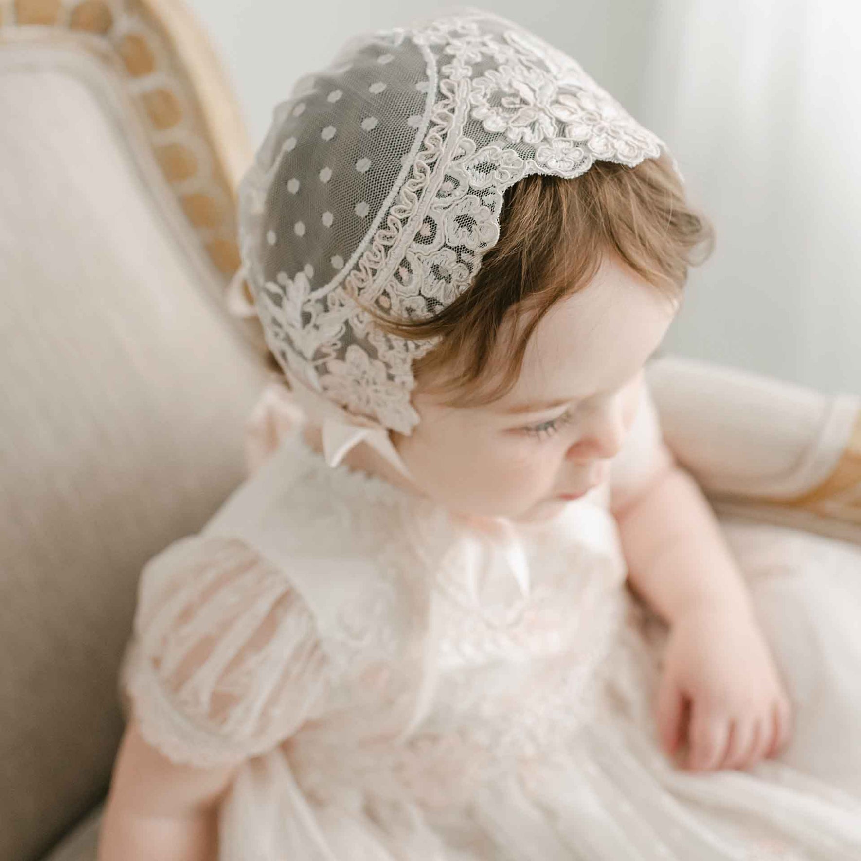 A young child wearing the Elizabeth Christening Gown & Bonnet, featuring exquisite handmade lace, sits on an upholstered chair. The softly lit setting accentuates the detailed patterns of the white ensemble as the child gazes to the side.