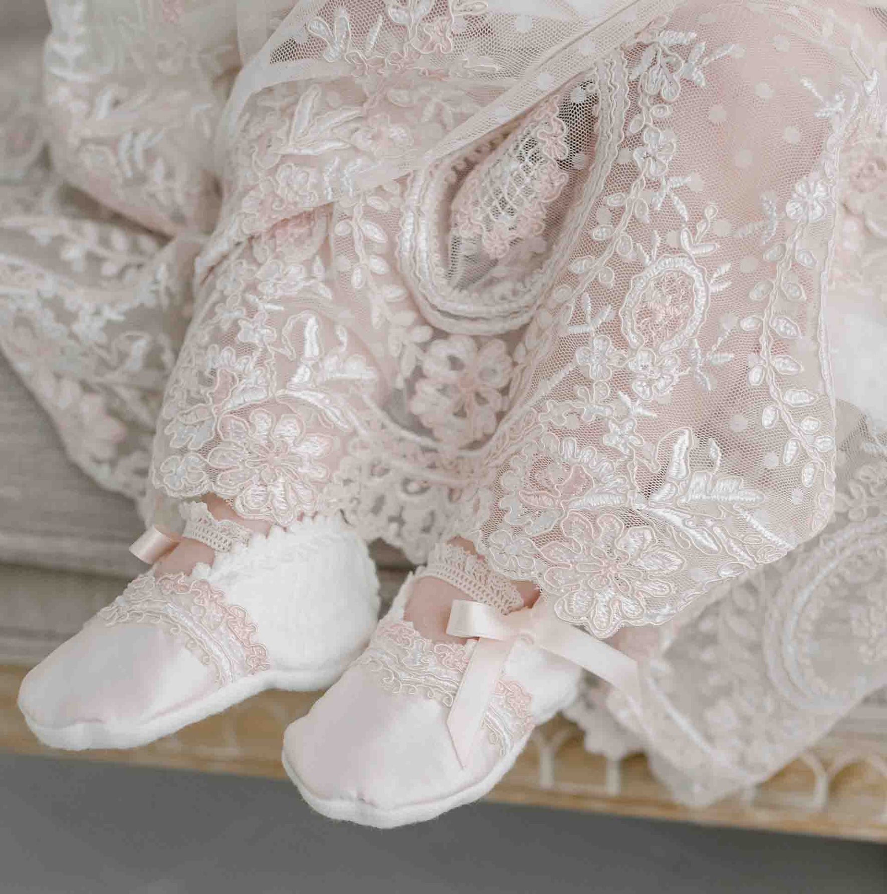 A close-up view showcases a baby wearing the Elizabeth Christening Gown & Bonnet, featuring intricately embroidered lace fabric and beautiful floral patterns. This handmade outfit includes white booties adorned with delicate lace and ribbon details, highlighting the elegance of silk Dupioni. The background is skillfully blurred for emphasis.