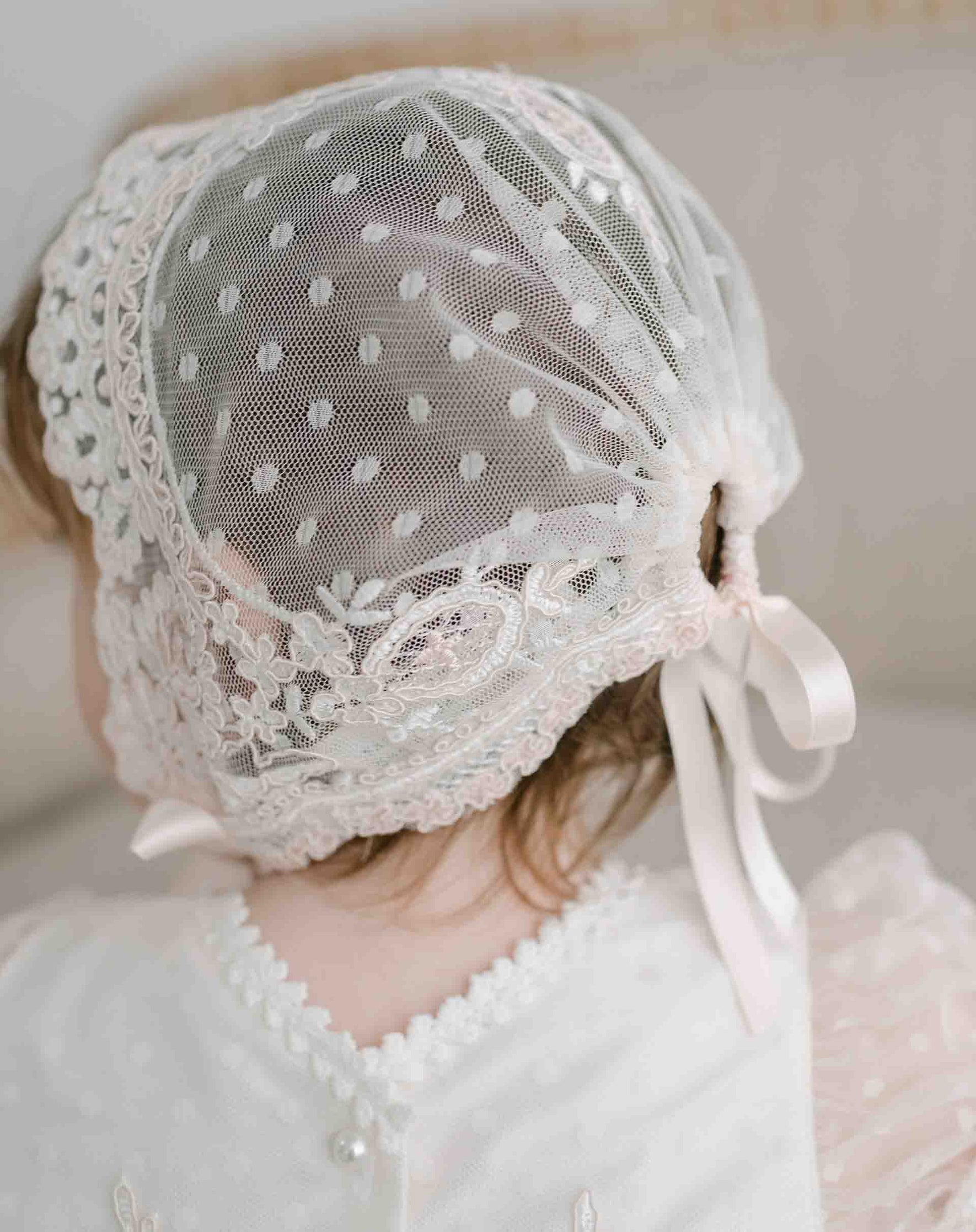 A child is seen from behind wearing the delicate Elizabeth Christening Gown & Bonnet, adorned with exquisite lace details and tied with a ribbon. The white baptism gown, crafted from silk Dupioni, features floral and polka dot patterns on the bonnet, highlighting its handmade elegance.