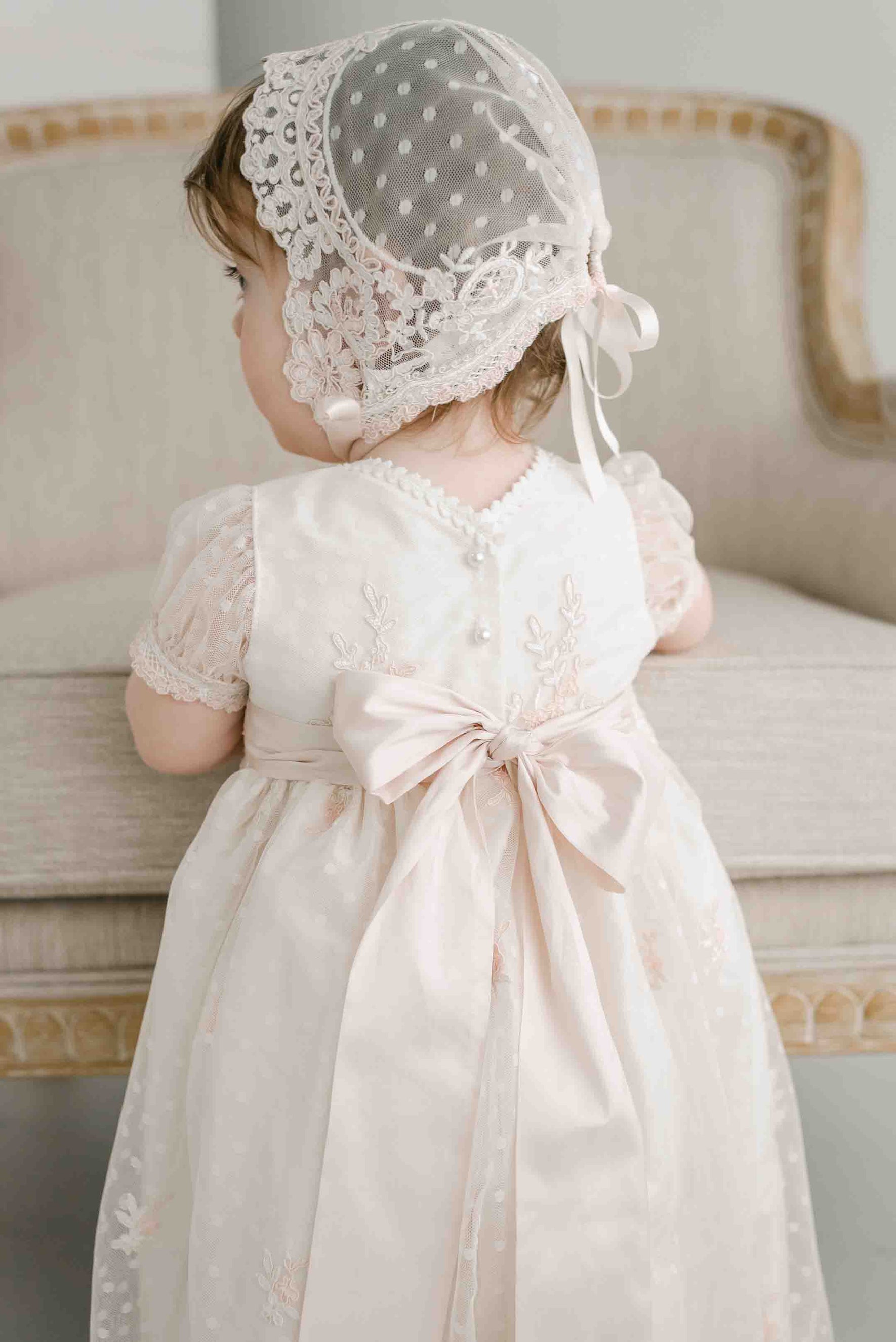 A young child sits on a light-colored upholstered chair, wearing the Elizabeth Christening Gown & Bonnet. The handmade gown is adorned with delicate lace and intricate floral patterns, and its embroidered cream silk Dupioni fabric, complete with a large bow on the back, showcases timeless beauty.