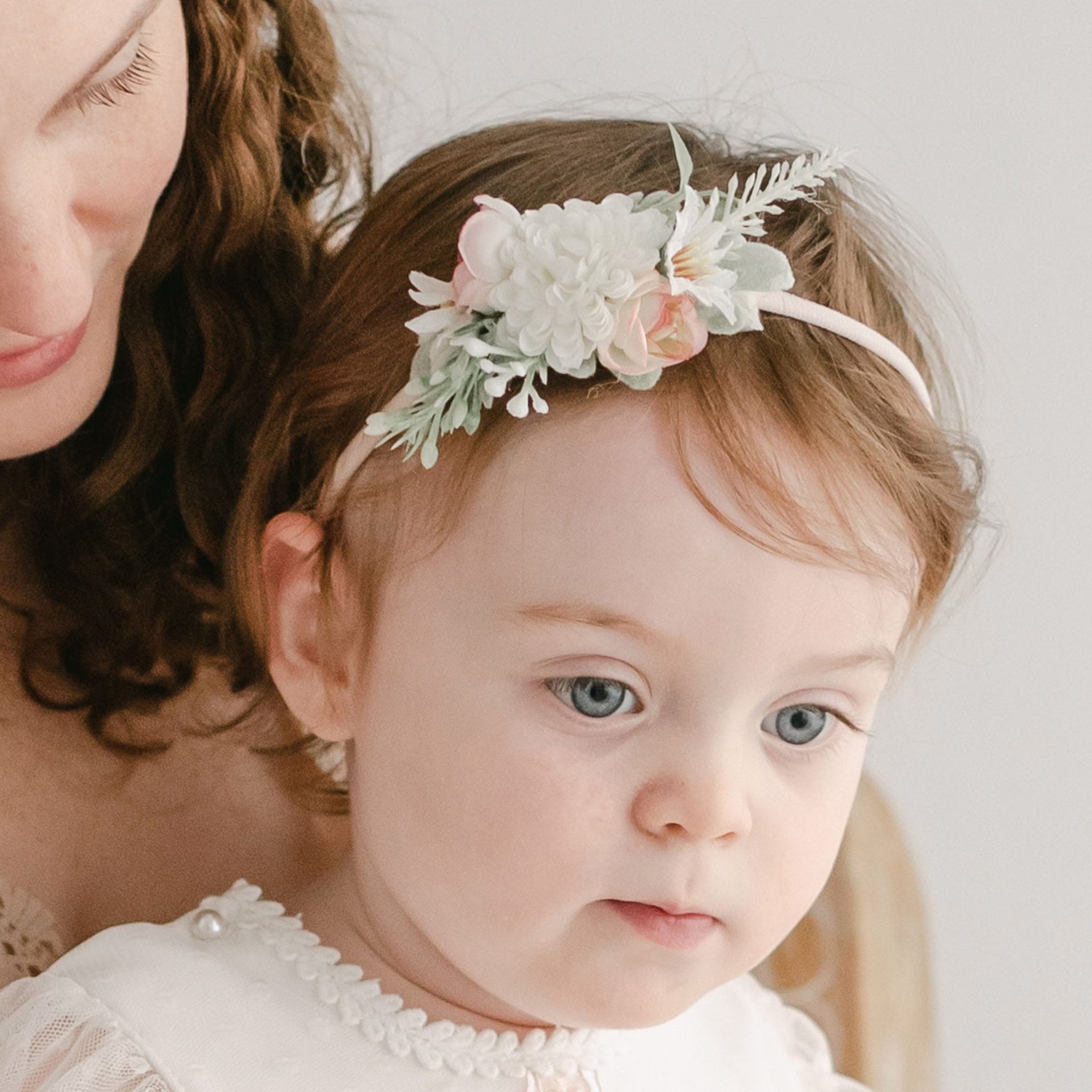 A young child with light brown hair, wearing the Elizabeth Flower Headband adorned with pink and cream flowers, sits close to someone with long, curly brown hair. The child is dressed in a light-colored outfit with delicate details and appears to be in a calm, neutral mood.