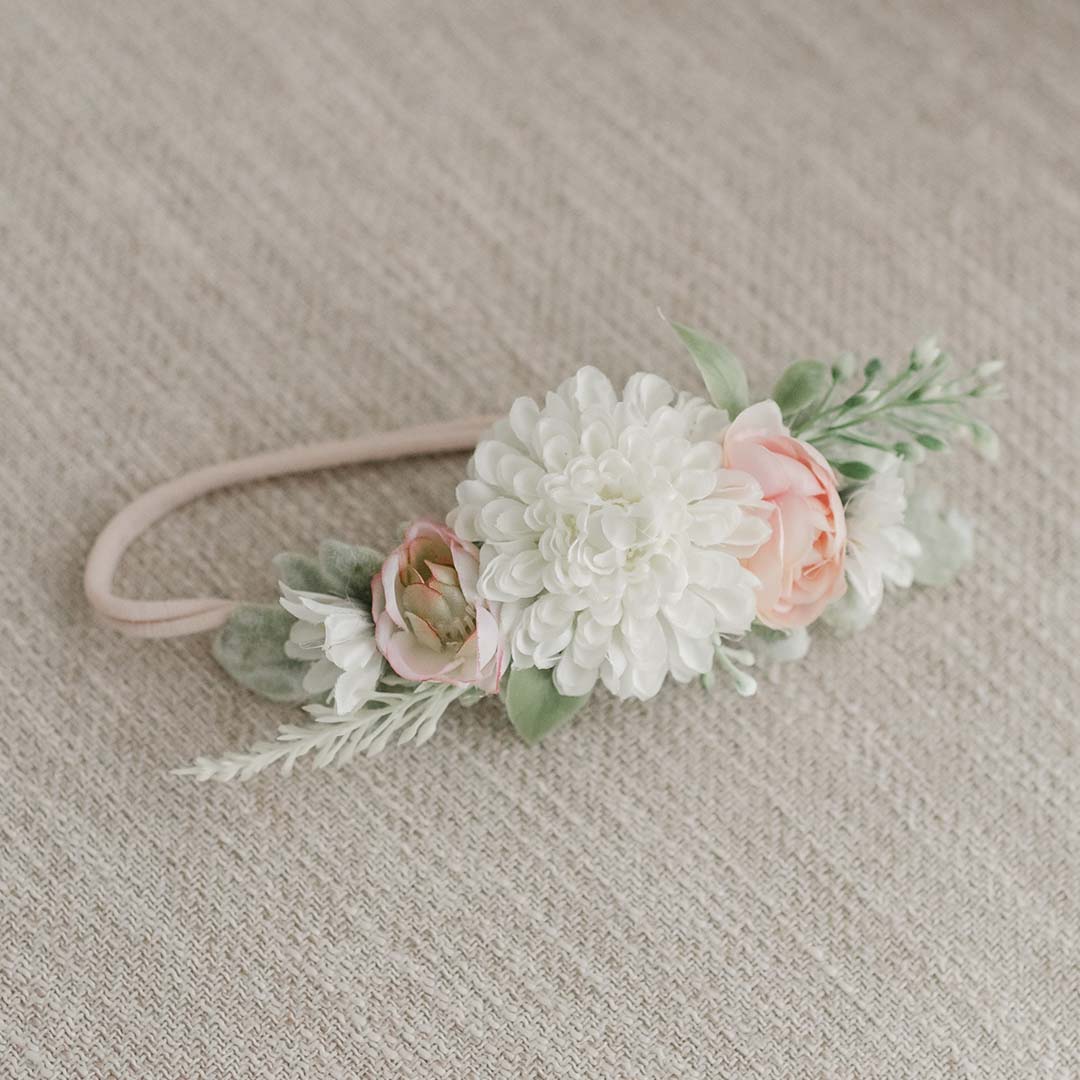 The Elizabeth Flower Headband is elegantly displayed on beige fabric, showcasing a large white chrysanthemum at its center. It is beautifully accented with two smaller pink roses, green leaves, and sprigs of foliage on either side. This delicate baby headband is adorned with charming pink and cream flowers.