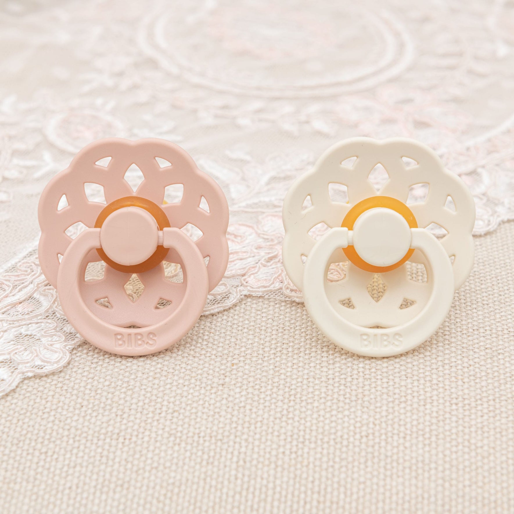 A pair of Elizabeth Pacifiers in blush and ivory rest on a lace-patterned surface. Crafted in Denmark, these natural rubber pacifiers boast round knobs and decorative cut-out designs on their shields, making them ideal for helping little ones self-soothe.
