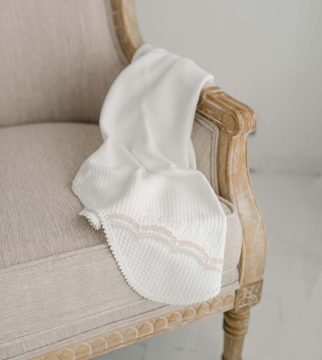 A beige armchair with a carved wooden armrest showcases the Elizabeth Personalized Blanket, elegantly draped over it. This white textured blanket, featuring delicate pink embroidery and scalloped edges, adds a touch of personalization against the light-colored, minimalist background.