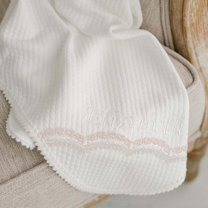 The Elizabeth Personalized Blanket, ideal for a christening outfit, elegantly adorns a beige chair. This textured white blanket features "Elizabeth" embroidered in white thread and is beautifully accented with a delicate pink lace trim along the edges.