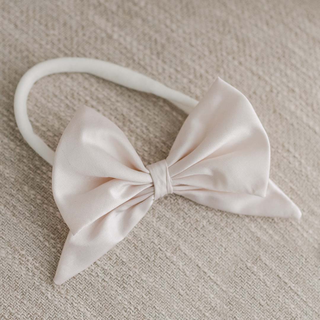 The Elizabeth Silk Bow Headband, featuring a delicate vintage pink champagne bow made of silk on a white elastic band, is elegantly displayed on a textured beige surface.