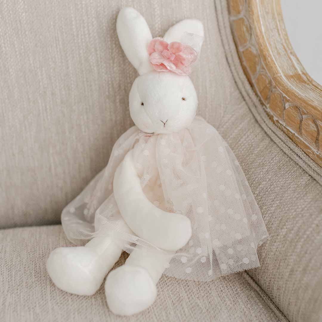 The Elizabeth Silly Bunny Buddy, a pacifier-holding bunny, cozily sits on a beige chair. This baby-safe companion features a white stuffed rabbit wearing a pink polka dot tutu and adorned with a charming pink flower on one ear, keeping your little one's pacifier always within reach.