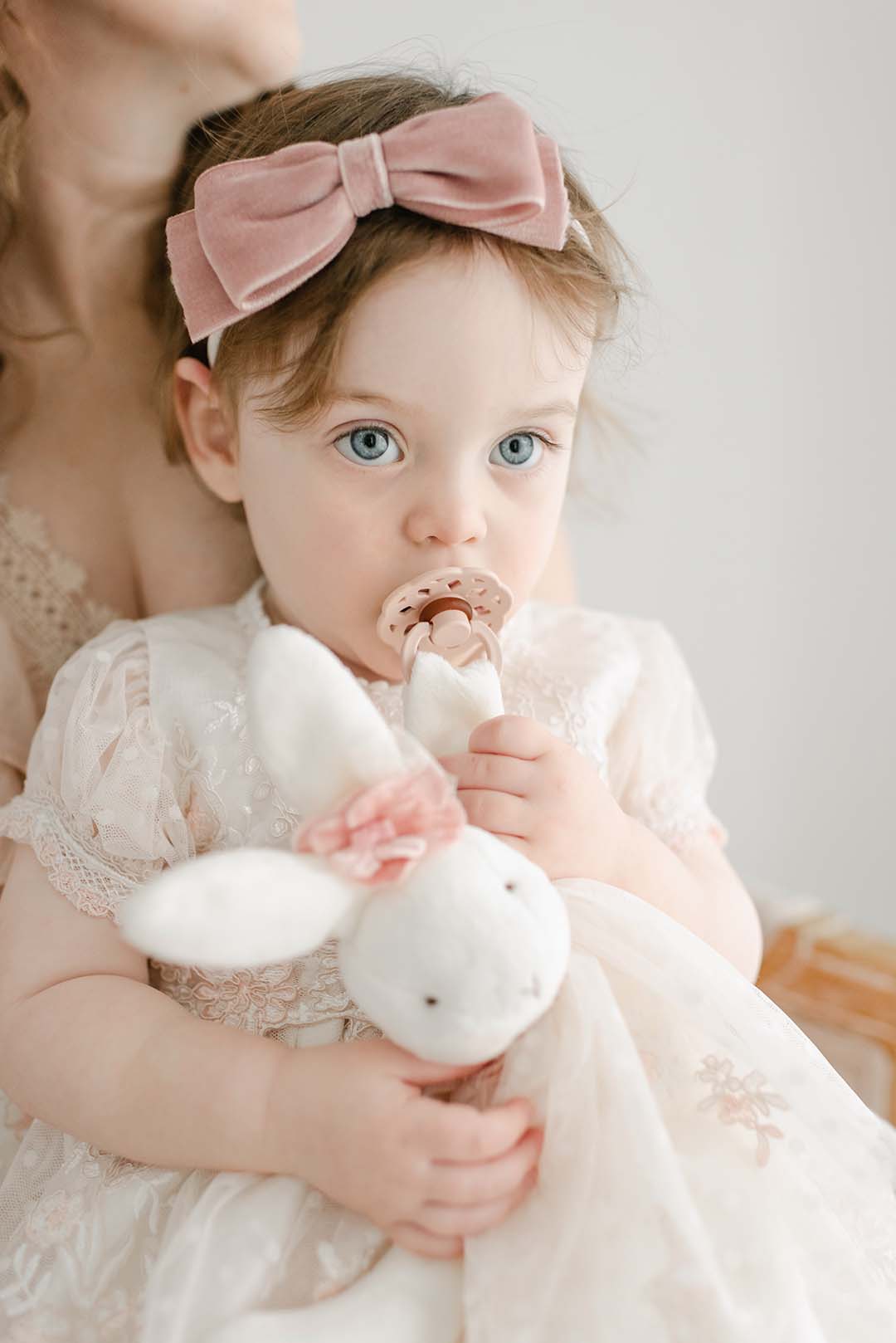 A toddler with a pink headband and pacifier holds the Elizabeth Silly Bunny Buddy, an adorable plush white rabbit that doubles as a pacifier holder. The child wears a light-colored dress with floral details, which is designed to be baby safe. An adult is partially visible in the background.