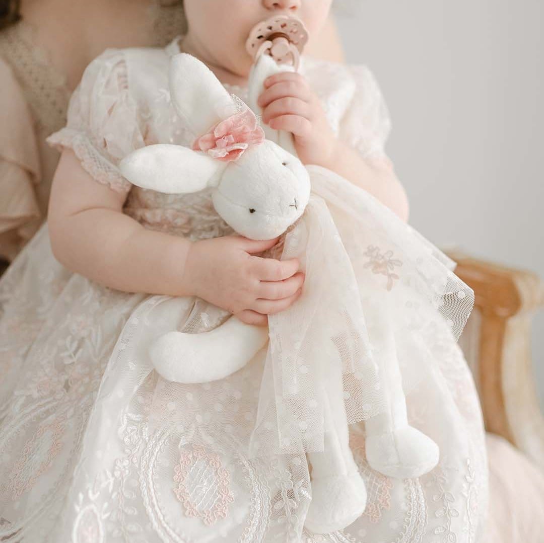 A toddler in a delicate pink lace dress holds the Elizabeth Silly Bunny Buddy, a stuffed white rabbit adorned with a pink bow and tulle skirt. Seated on a wooden chair, the child uses this baby-safe buddy as a pacifier holder attached to a cute clip.