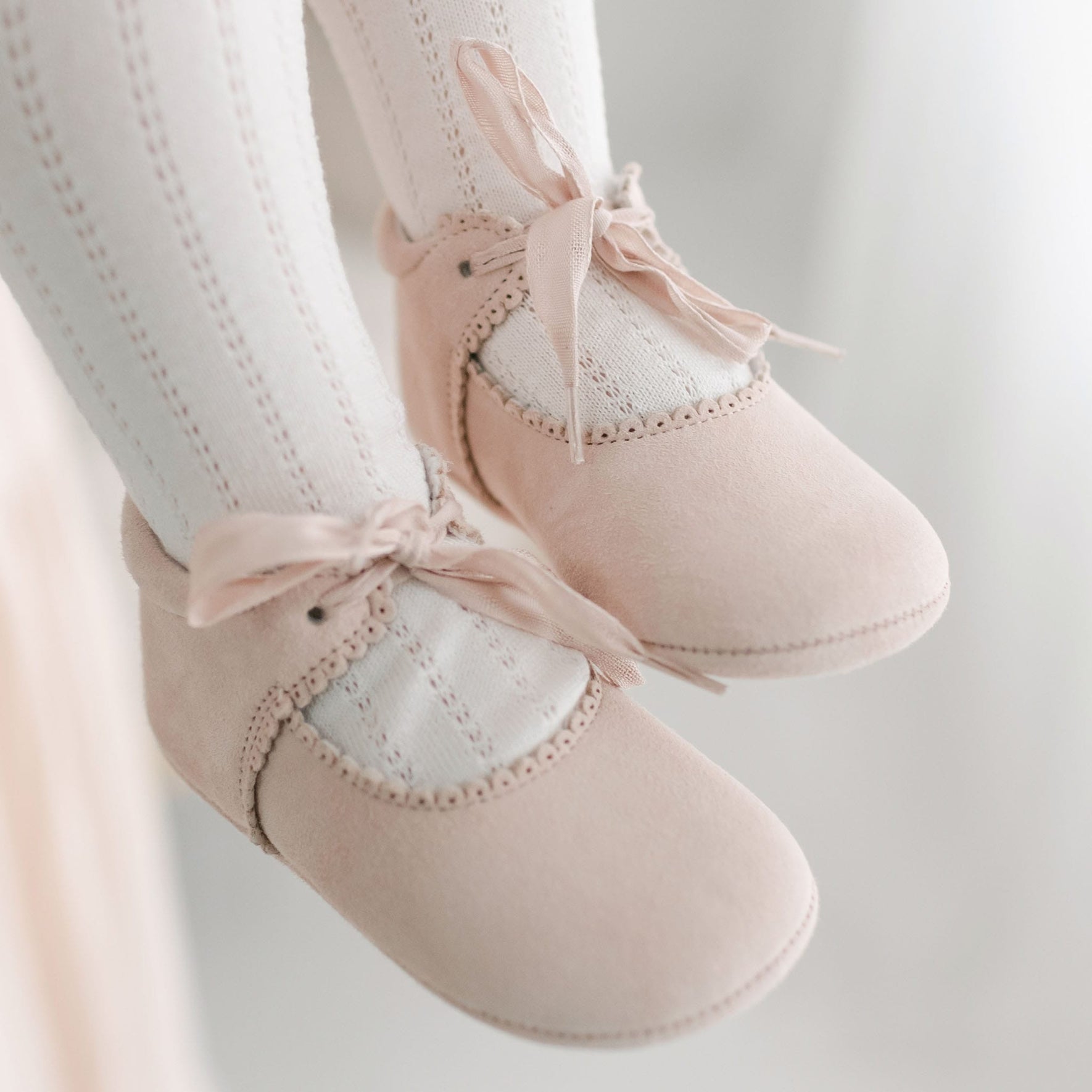 Close-up of a pair of baby's feet wearing white knitted socks and Elizabeth Suede Tie Mary Janes with ribbon ties. The blush suede shoes feature decorative scalloped edges, and the background is blurred.