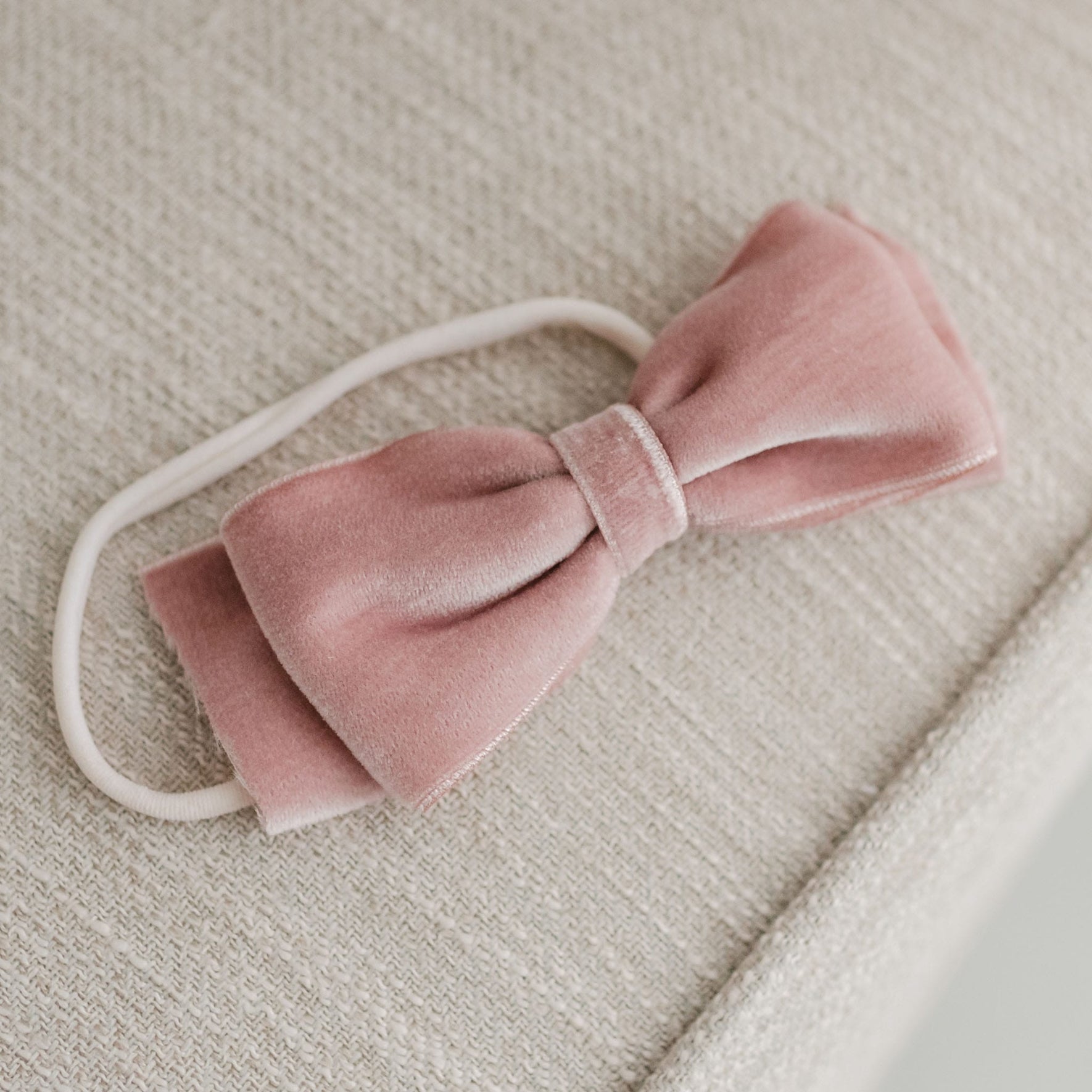 The Elizabeth Velvet Bow Headband, in a pink velvet finish, rests on a beige textured surface. It features a simple knot design for a comfortable fit and is attached to a white elastic band.