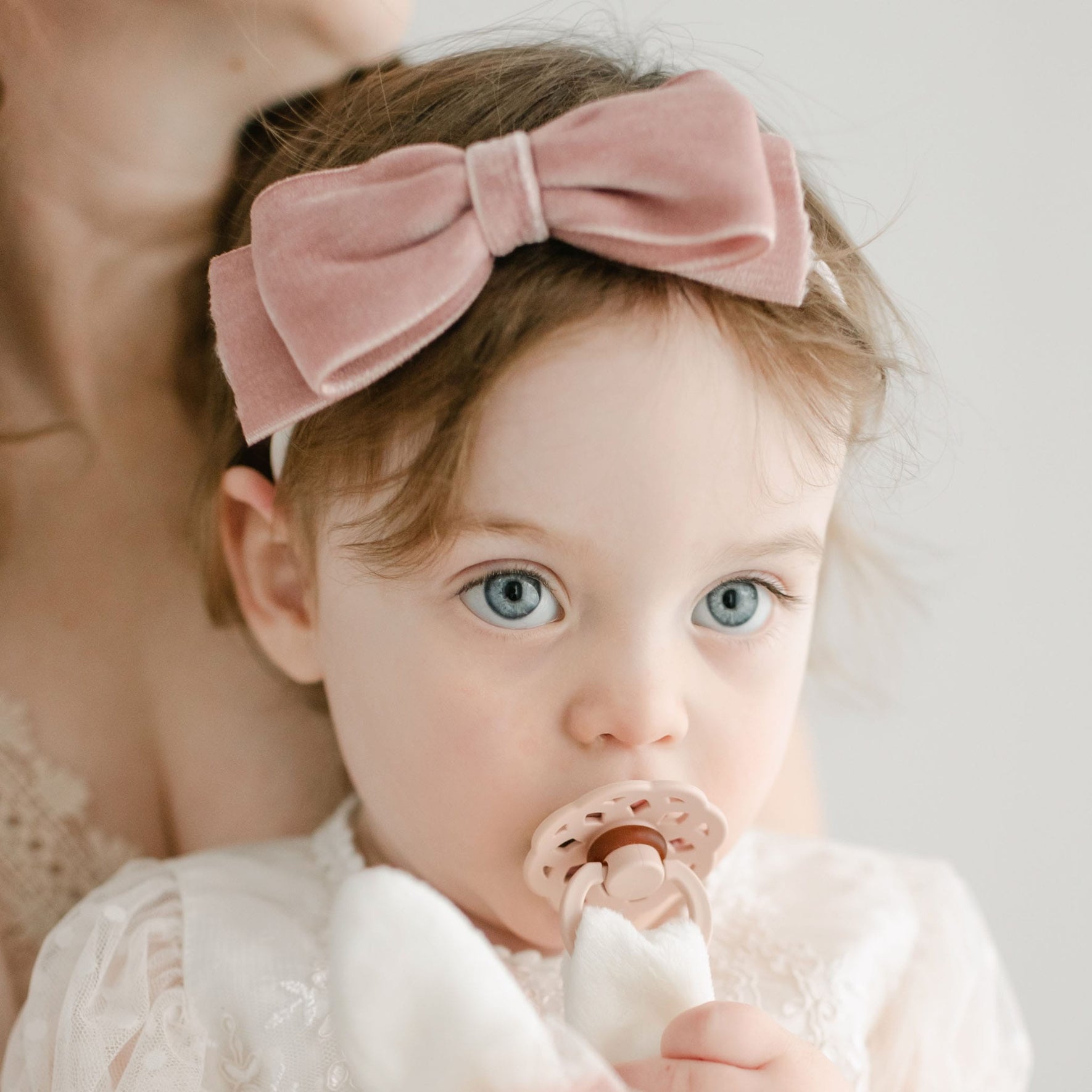 A young child with light brown hair is wearing the Elizabeth Velvet Bow Headband, which is handmade in the USA. The child, appearing cozy in a white outfit, holds a pacifier in their mouth. Part of an adult's face and shoulder can be seen in the background, contributing to the serene scene.