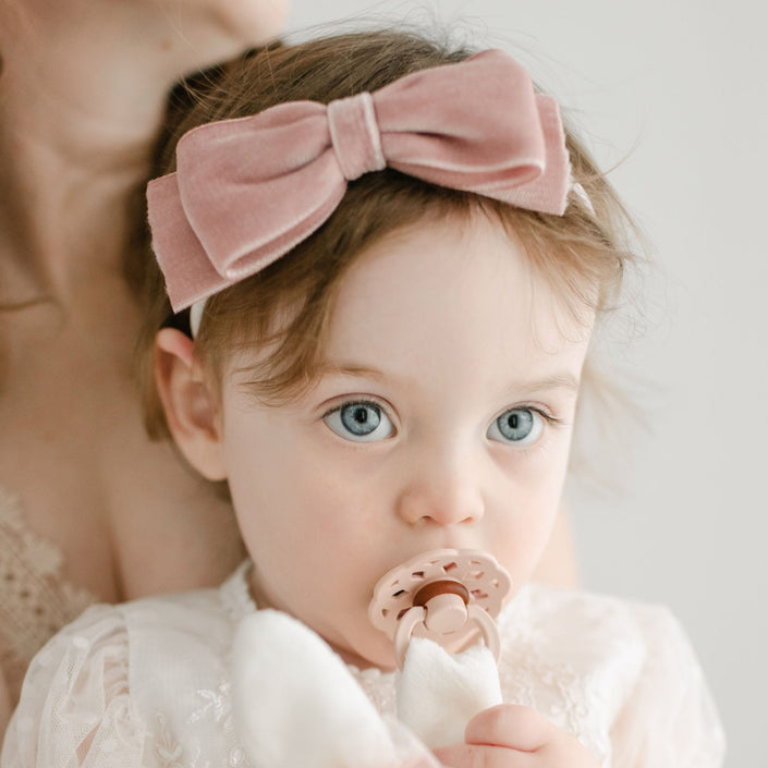 A young child with light brown hair is wearing the Elizabeth Velvet Bow Headband, which is handmade in the USA. The child, appearing cozy in a white outfit, holds a pacifier in their mouth. Part of an adult's face and shoulder can be seen in the background, contributing to the serene scene.