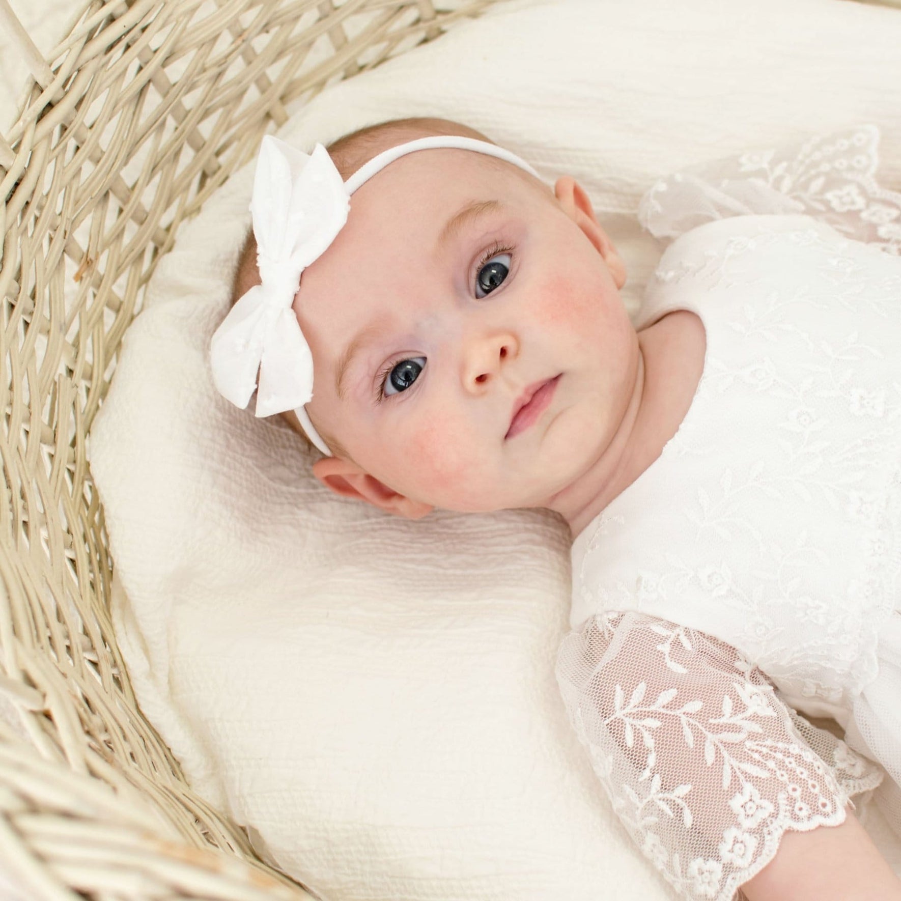 A newborn baby girl gazes into the camera with wide eyes, wearing the Ella Baby Girl Headband and matching romper dress.