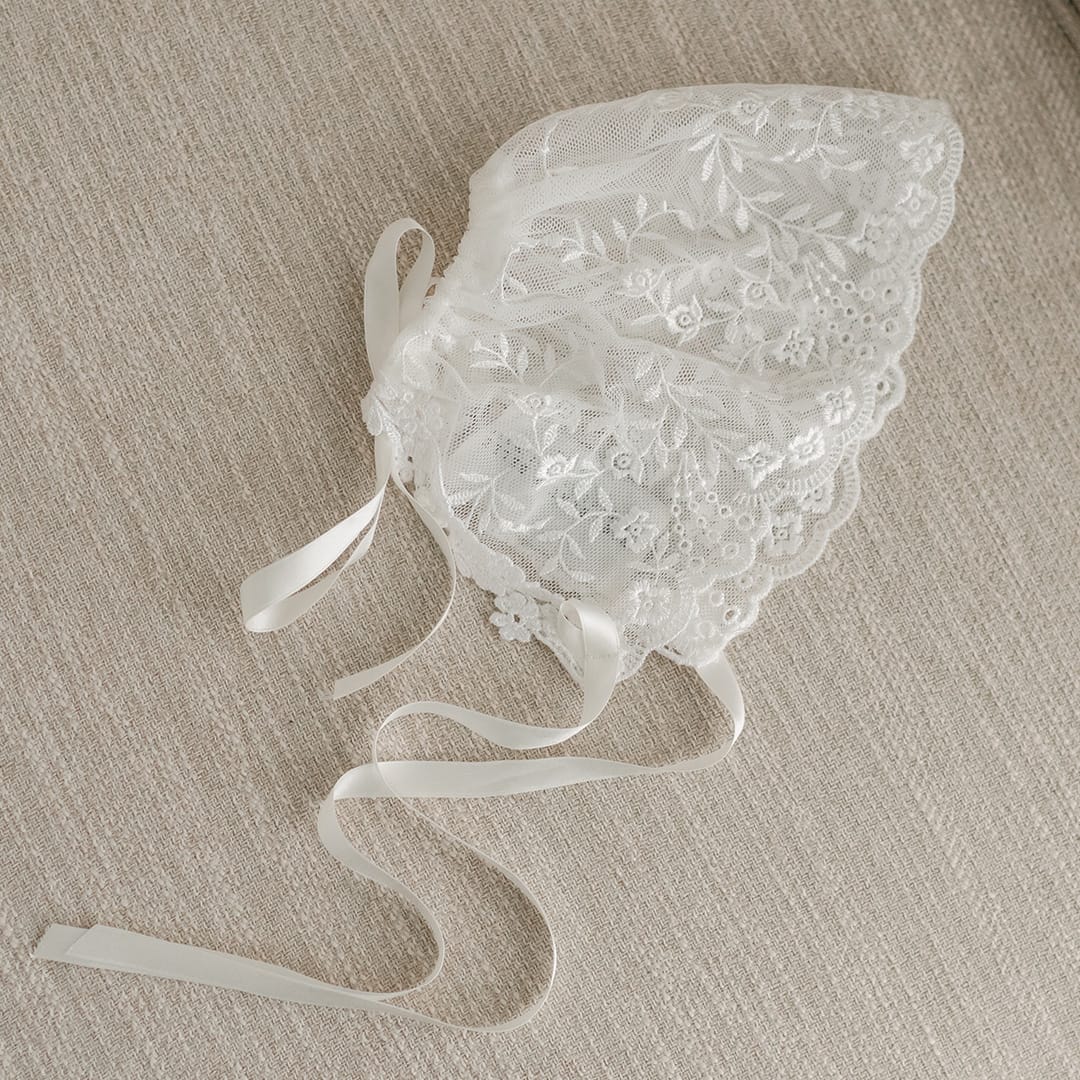 A flat lay photo of the boutique Ella Lace Bonnet on a beige fabric chair.