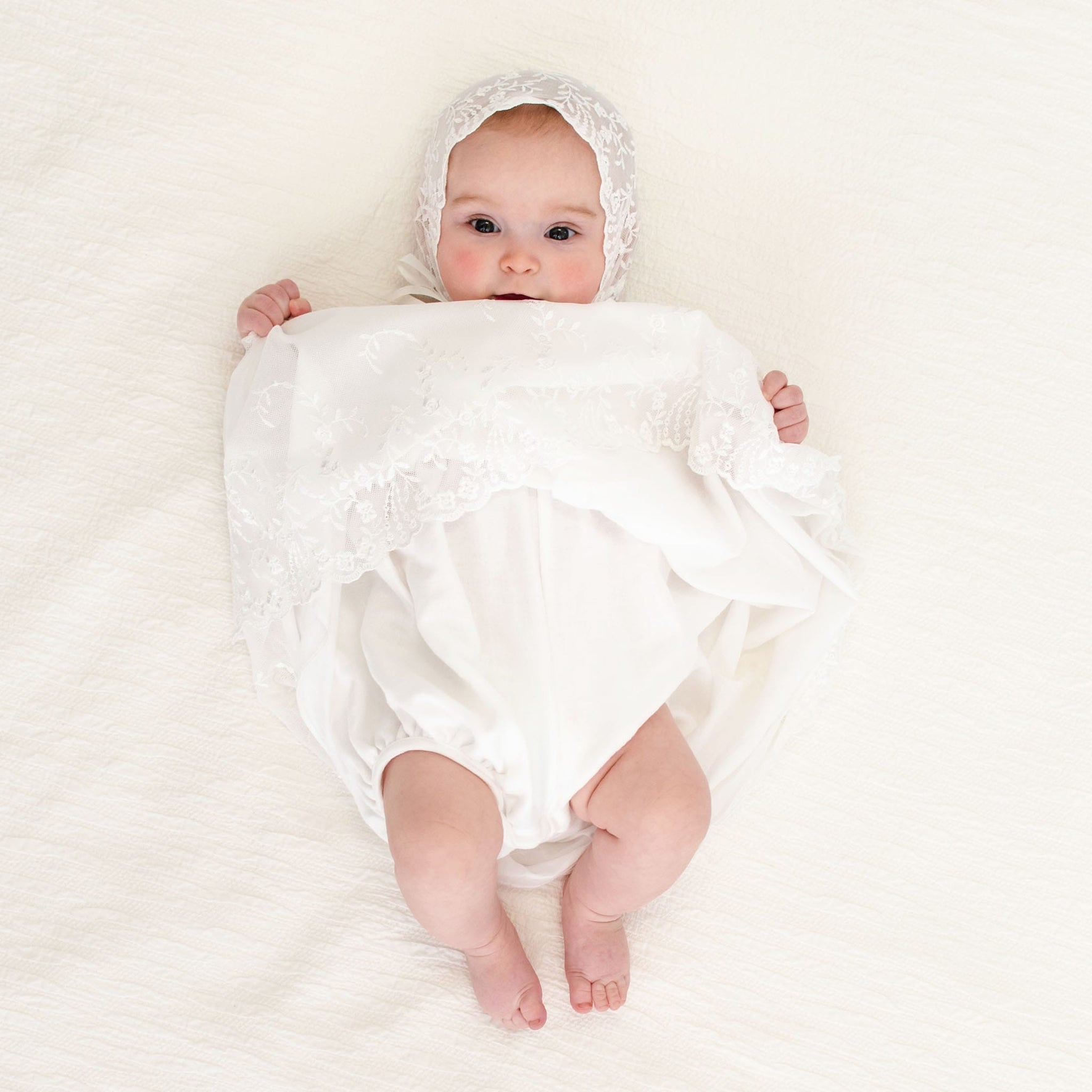 A baby girl holding up her skirt showcasing the detail under lace hem of the Ella lace baptism dress.