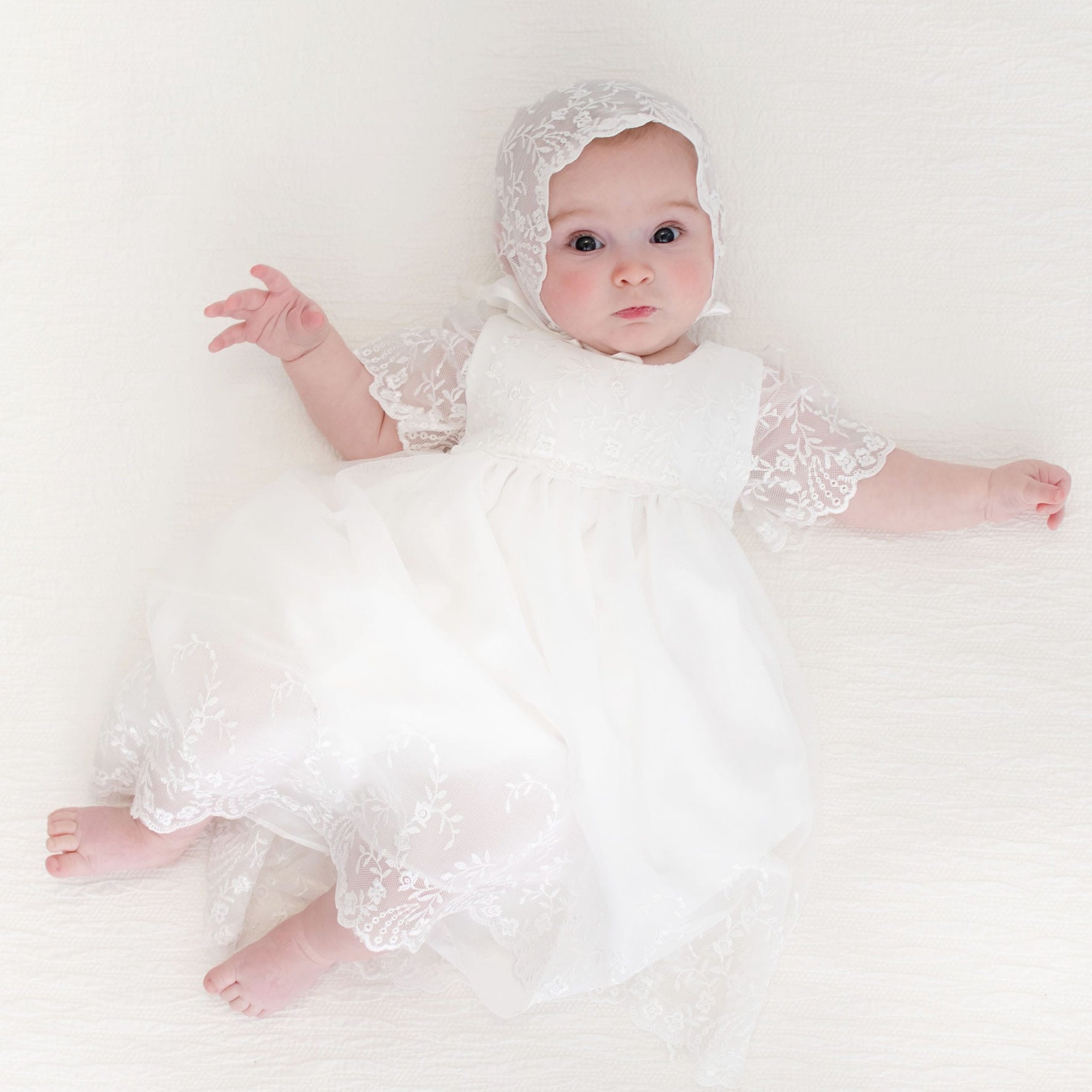 Baby girl dressed in the Ella baptsim dress and Ella Lace Bonnet, gazing curiously at the camera with open arms.