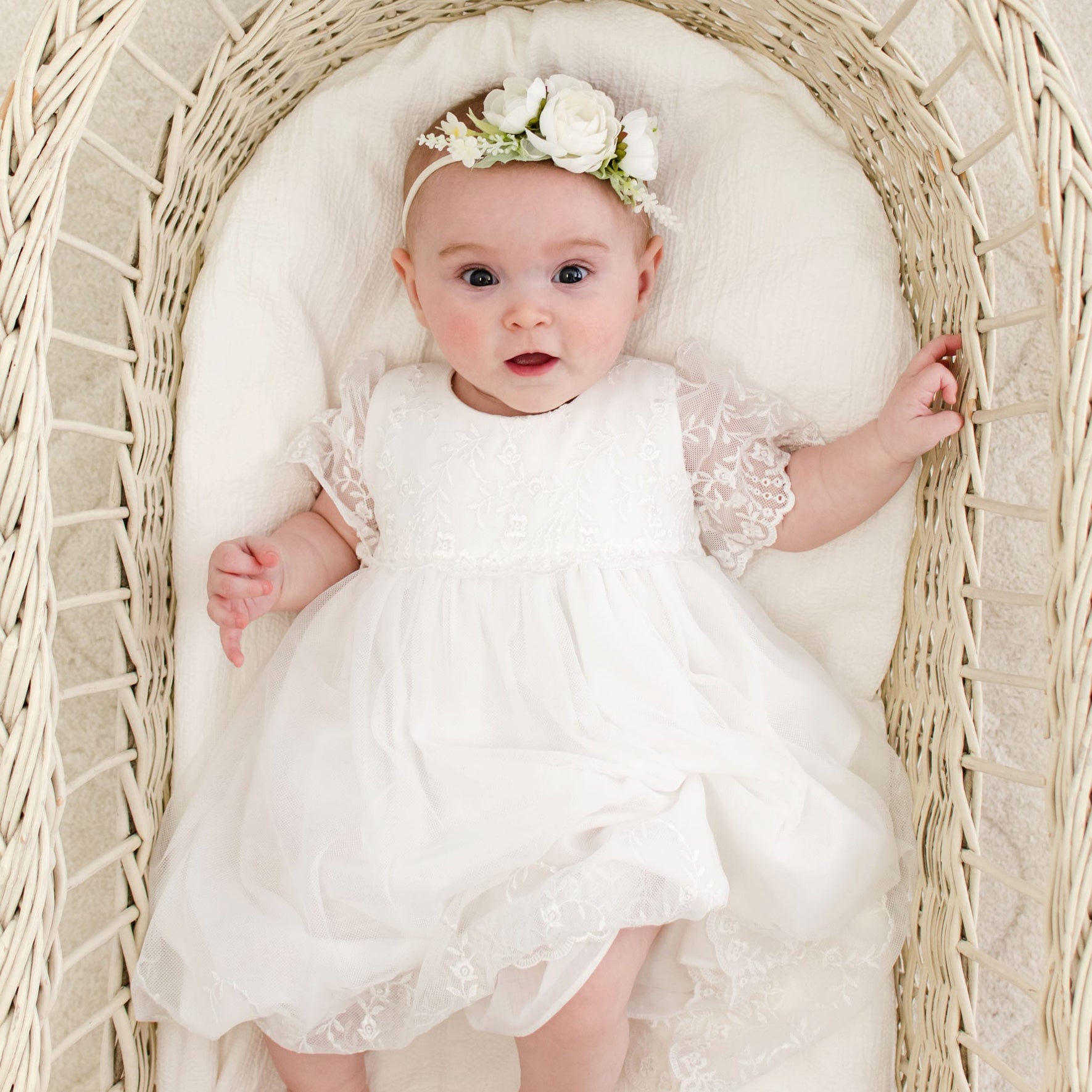 A baby girl wearing a vintage-inspired Ella baptism dress and flower headband lies in a wicker bassinet.