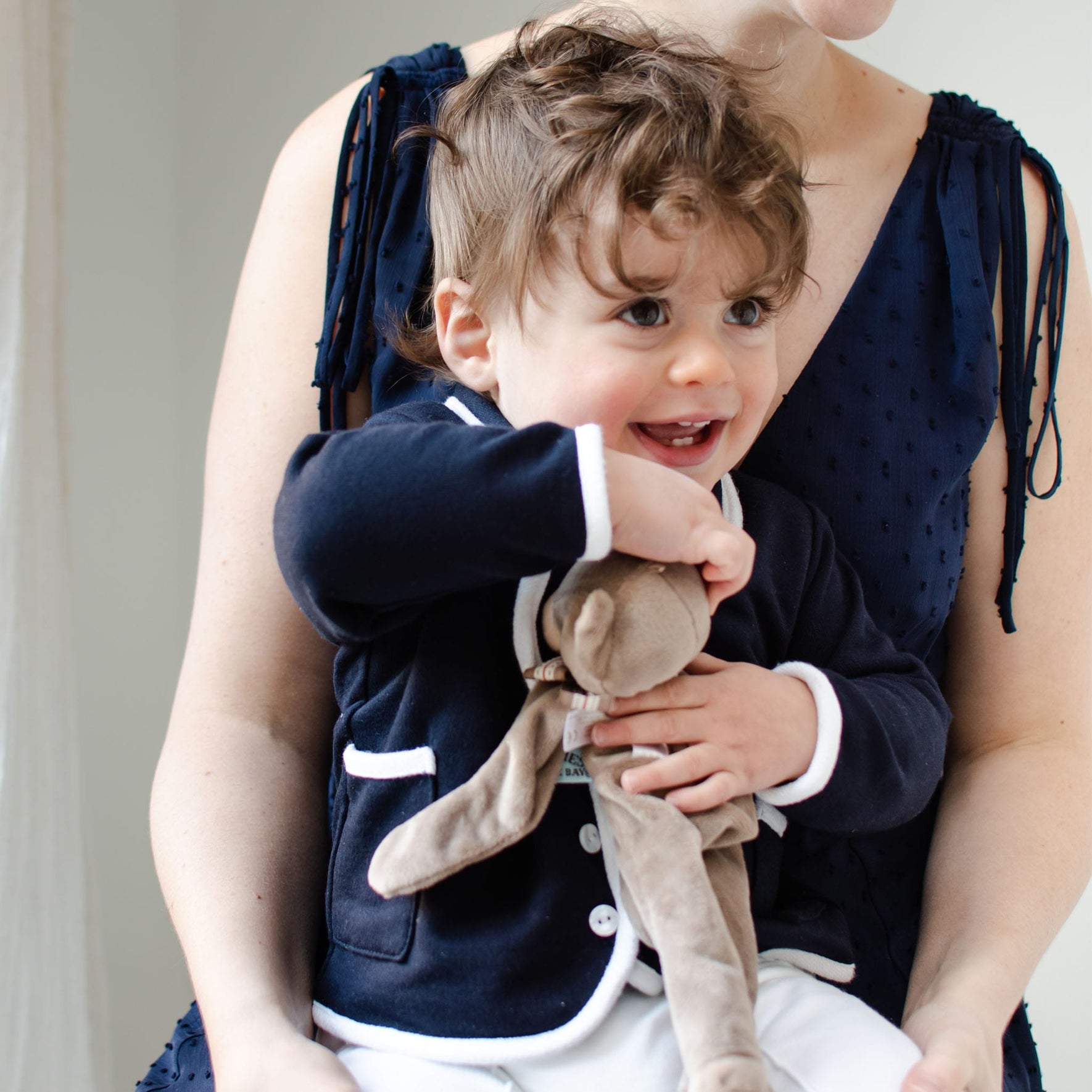 A toddler with curly hair, wearing an Elliott Jacket crafted from soft French terry cotton, sits on a woman's lap. Clutching a handmade stuffed animal, the child beams with joy. The woman complements his style in a sleeveless navy dress.
