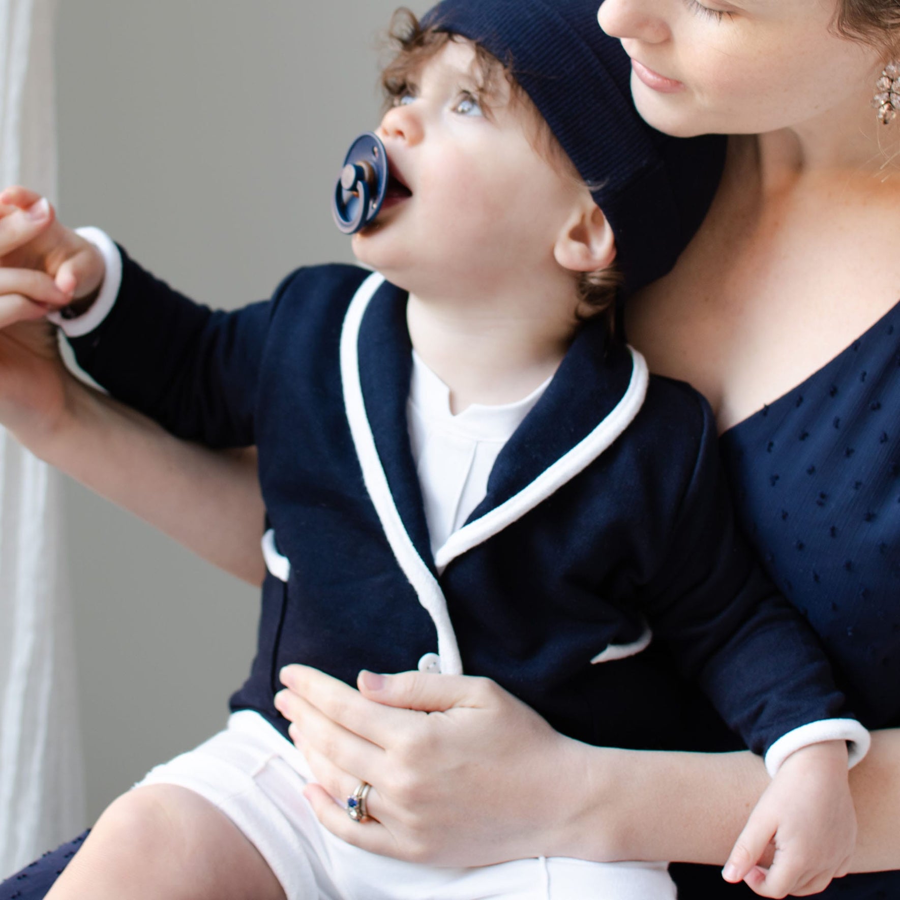A young child dressed in a handmade navy blue Elliott Jacket with white trim and a matching hat sits on an adult's lap. The child, who has a pacifier and is gazing upward, is held by the adult wearing a blue dress, who gently holds the child's hand.