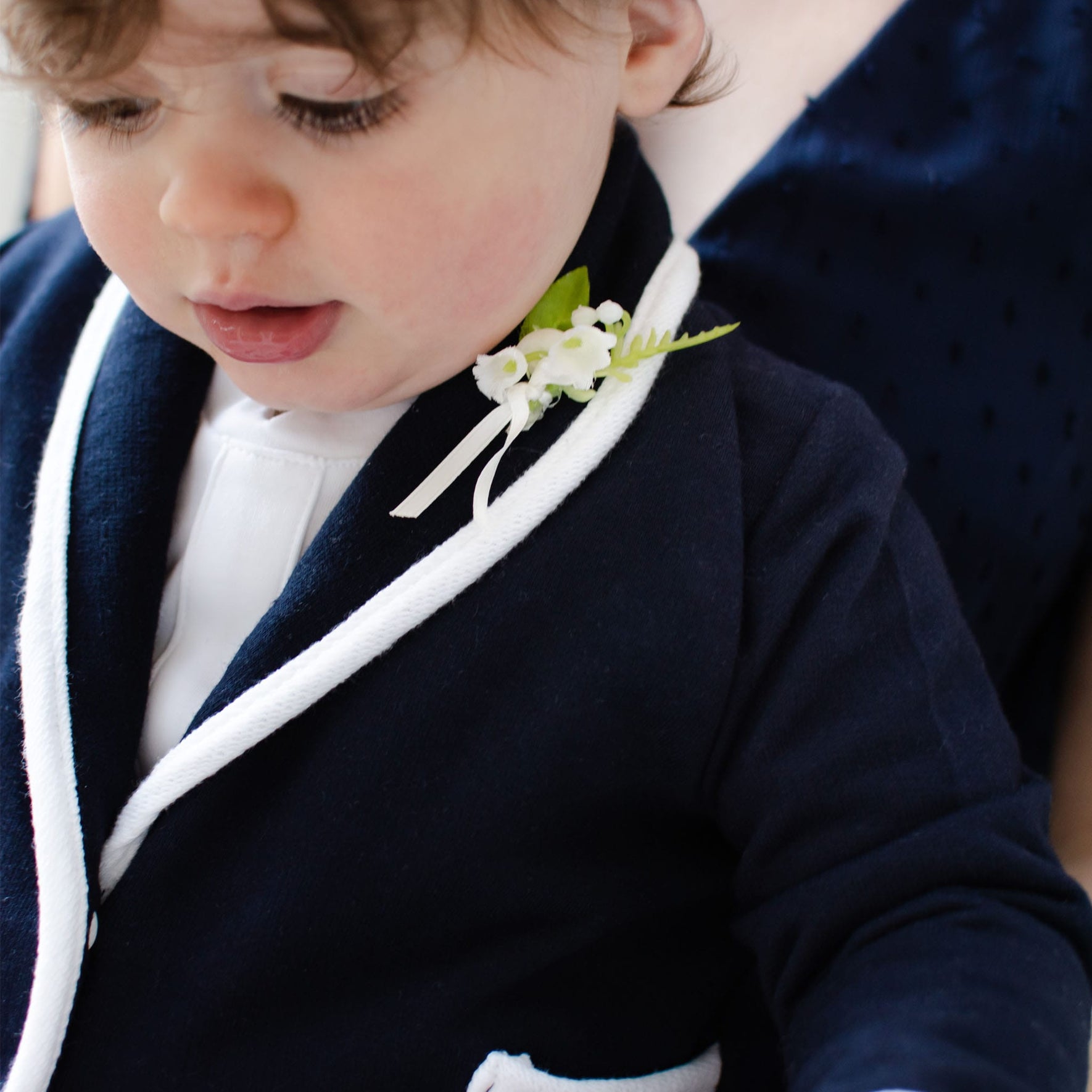 A toddler wearing an Elliott Jacket in navy, featuring white trim and a lily of the valley boutonniere, gazes downwards. The handmade piece is crafted from soft French terry cotton, with a blurred figure of an adult in a dark outfit appearing in the background.