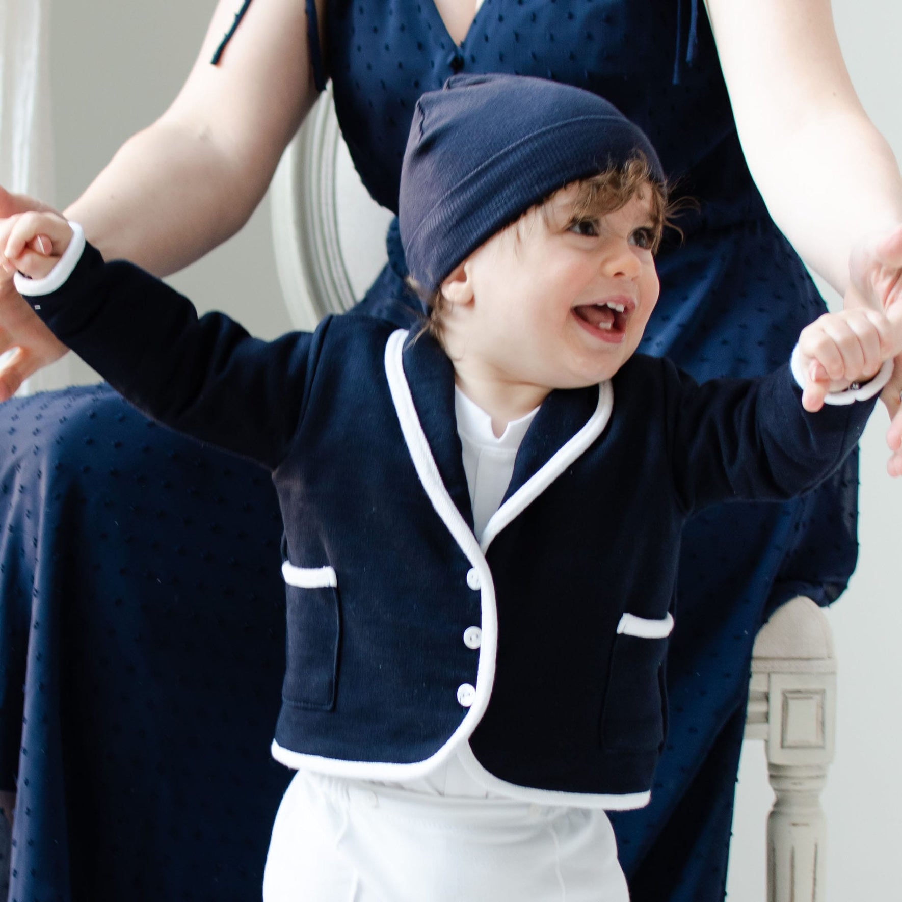 A toddler wearing the Elliott Jacket, which is a navy French terry cotton jacket with white trim, and a matching hat smiles with arms outstretched. An adult in a navy dress is seated behind, holding the child's hands.