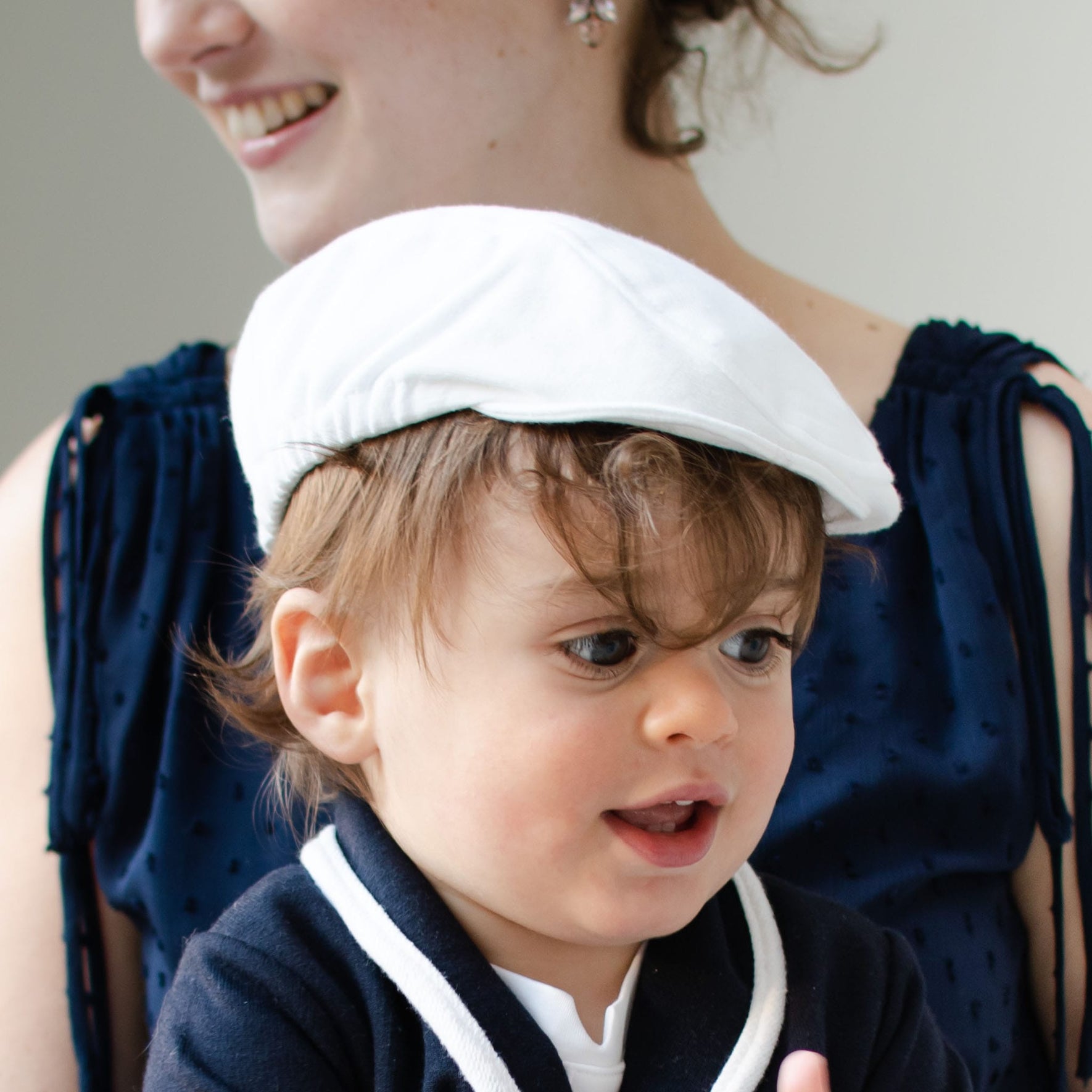 A young child sporting an Elliott Newsboy Cap is being held by an adult. The child, with brown hair, is dressed in a navy blue and white ensemble crafted from soft French terry cotton. The adult is partly visible, clad in a navy blue top.