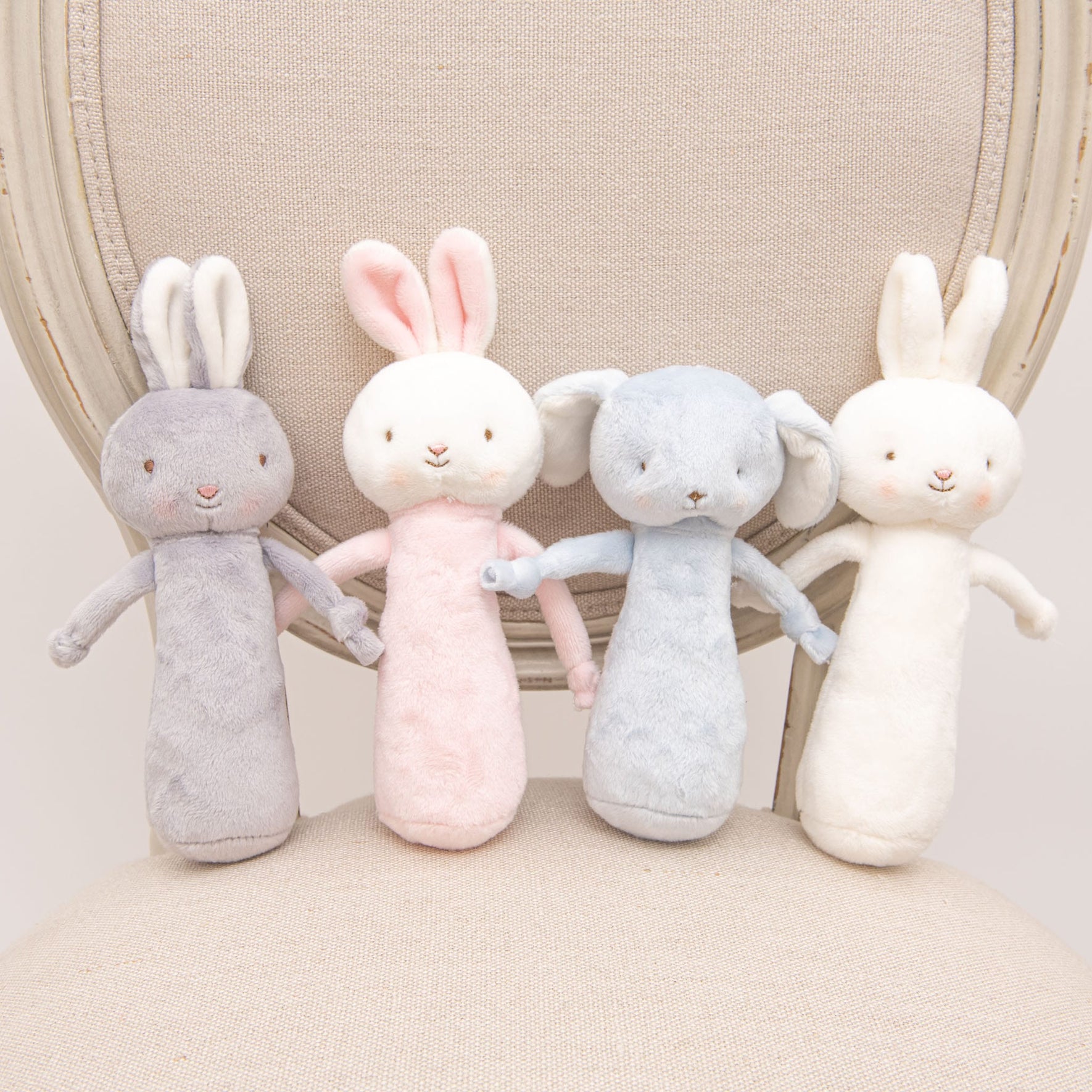 Four Friendly Chime Rattles in pastel colors—gray, pink, blue, and cream—are seated on a cushioned chair. Each baby-safe rattle features floppy ears and soft embroidered details, perfect for gentle cuddles.