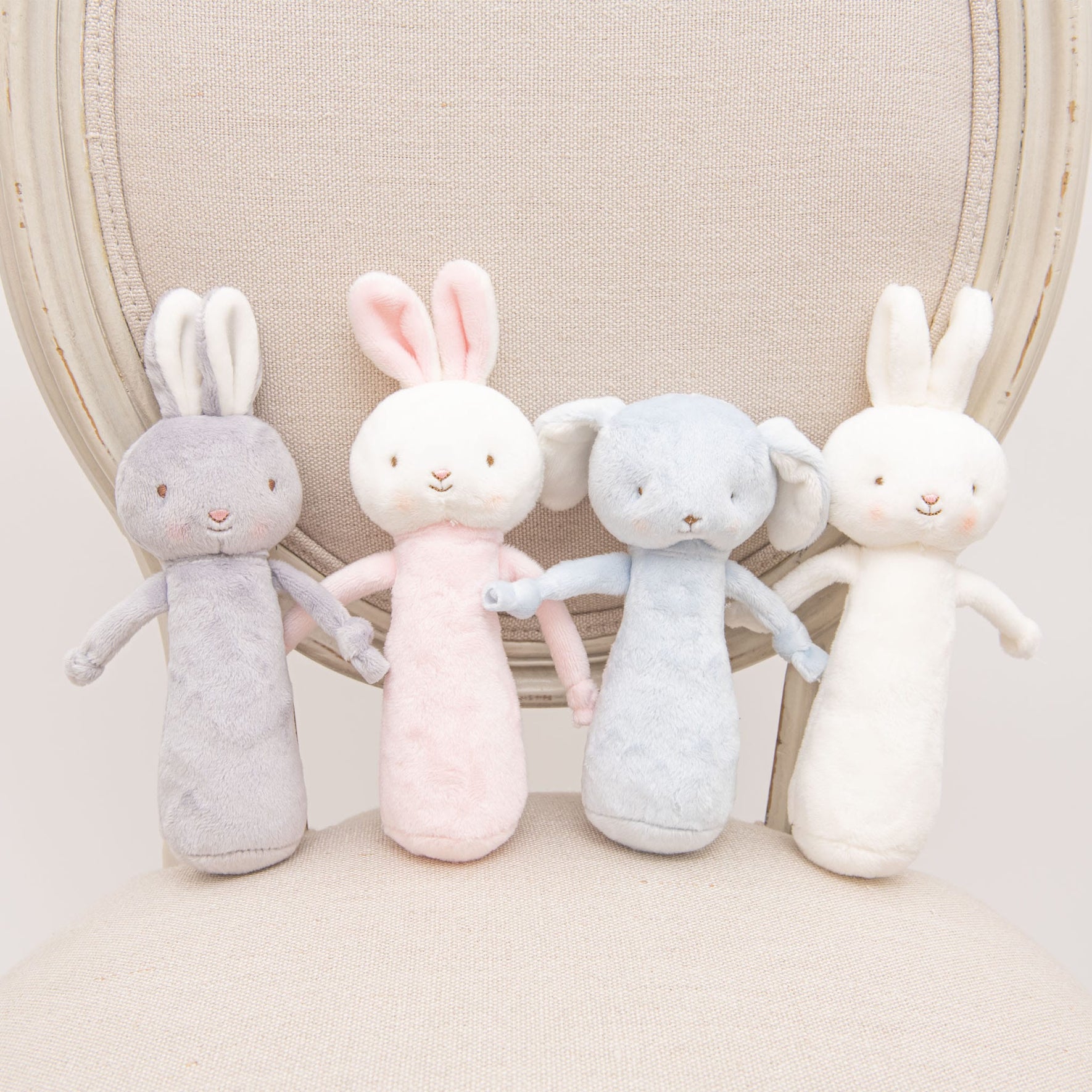 Four Friendly Chime Rattle plush toys in pastel colors—gray, pink, blue, and white—are sitting on a light-colored upholstered chair. Each baby-safe rabbit features soft embroidery and holds the paw of the next, forming a connected line.
