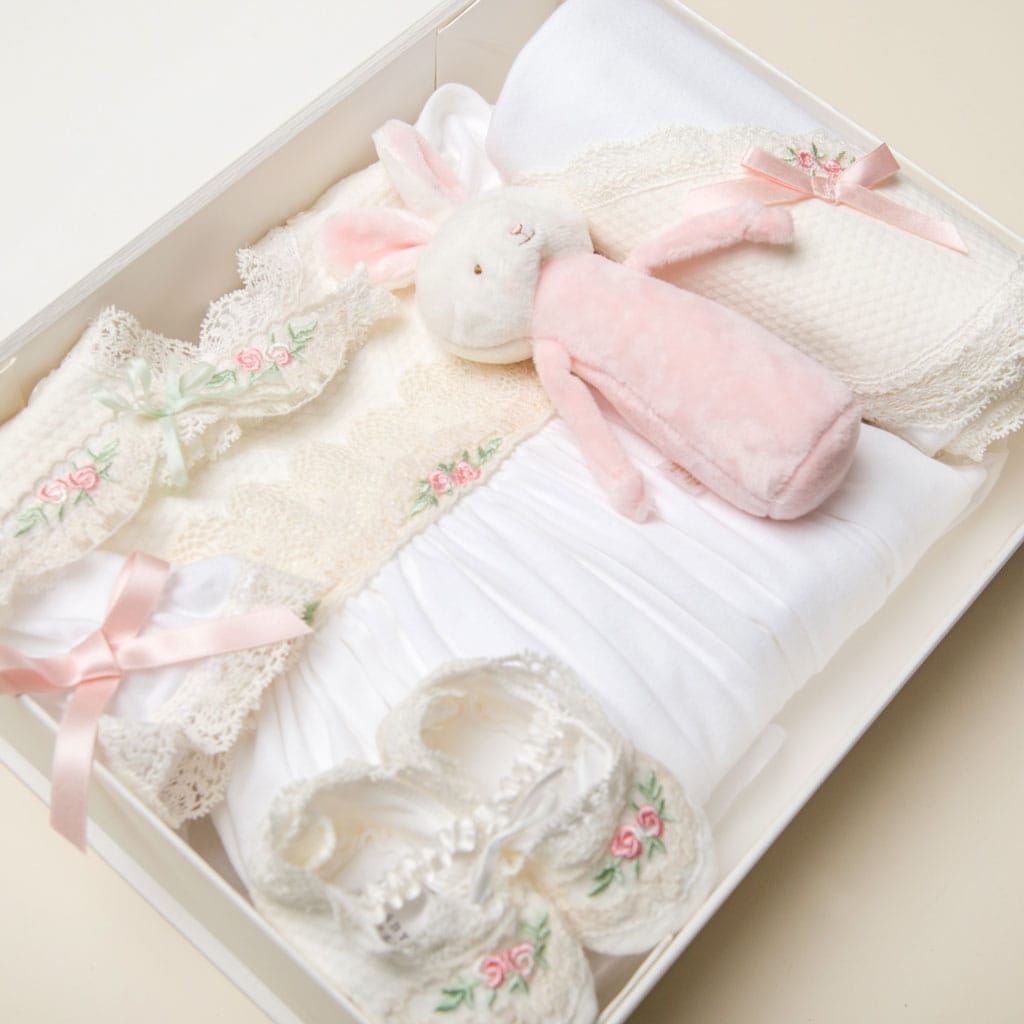 The beautifully arranged Ivory Gift Box from Baby Beau & Belle includes delightful baby items like a pink plush bunny toy, white knitted booties with floral embroidery, and a white lace trim blanket. Pink ribbons and floral details adorn the collection, making it perfect for cherishing.