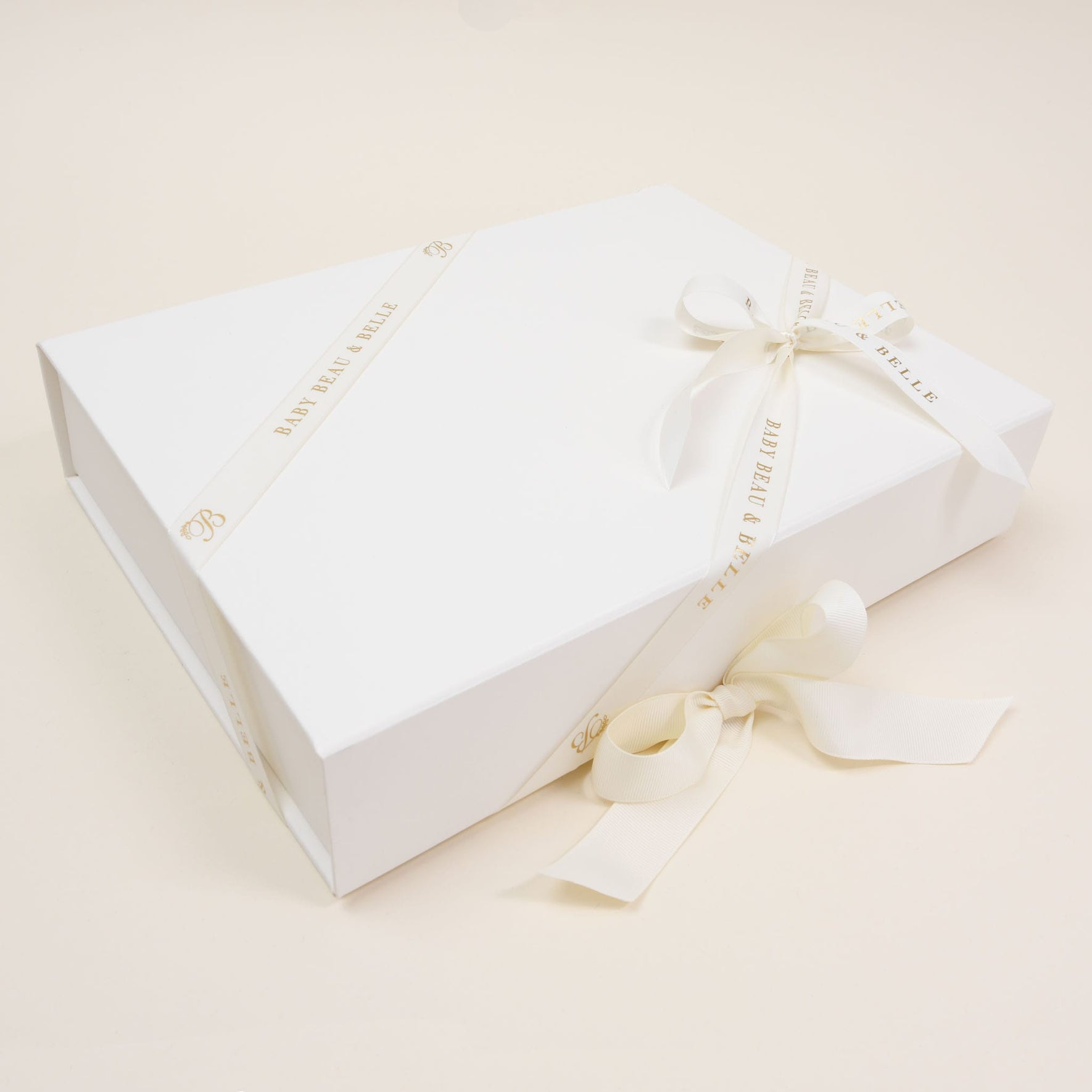 A large rectangular ivory gift box from Baby Beau & Belle, known as the Gift Wrap | Ivory Gift Box, is adorned with a satin cream ribbon elegantly tied into a bow. This exquisite keepsake showcases subtle gold text along the ribbon's edge, set against a plain off-white background.