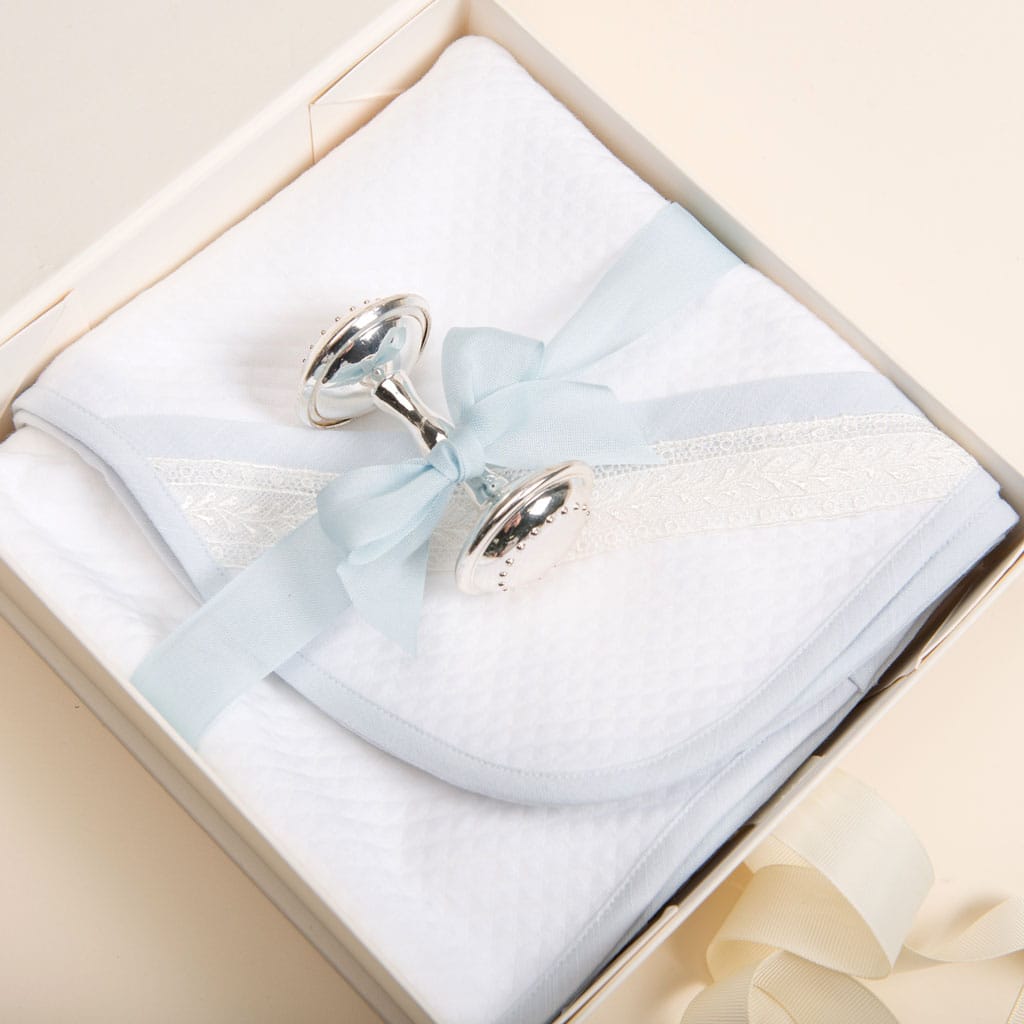 Inside the Ivory Gift Box, a neatly folded white blanket showcasing a textured pattern and embellished with a light blue ribbon and lace rests elegantly. Atop the blanket is a silver rattle adorned with a blue bow—an exquisite keepsake from Baby Beau & Belle. The box features an interior that is cream-colored.