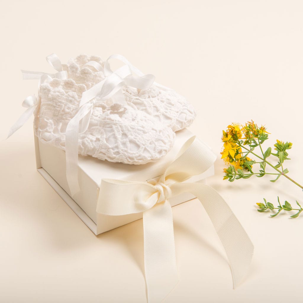 A pair of white lace baby shoes from Baby Beau & Belle, featuring satin ribbons, rest on an Ivory Gift Box from the Gift Wrap collection, adorned with a broad ribbon. A delicate sprig of yellow flowers is positioned next to this cherished keepsake on a soft beige background.