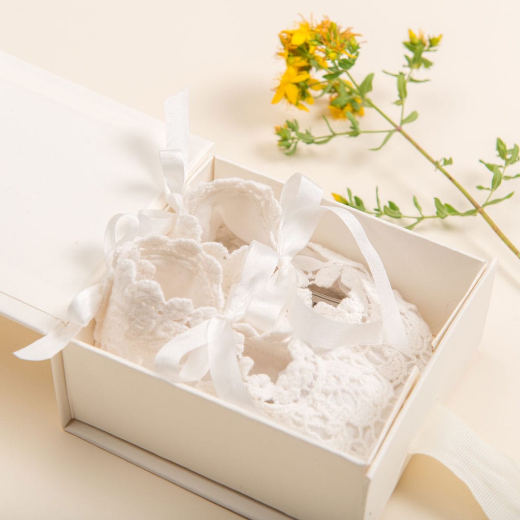 A white lace baby bonnet with ribbons by Baby Beau & Belle is elegantly placed inside a Mini Gift Box with Bow. Beside the box is a small cluster of yellow flowers on a light beige background, making it a perfect keepsake for cherished memories.
