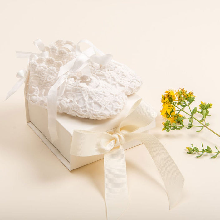 A pair of white crocheted baby shoes from Baby Beau & Belle, featuring ribbon ties, is elegantly displayed on a cream-colored Mini Gift Box with Bow. Beside this keepsake, a small yellow flower rests on a pale background.