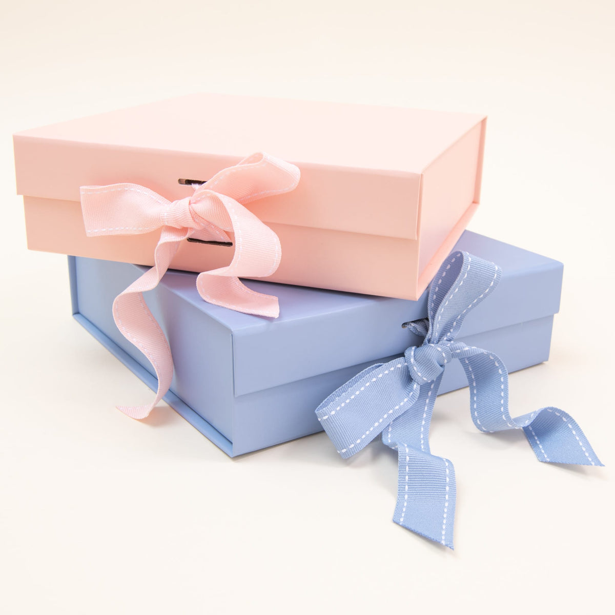Two gift boxes from *Gift Wrap | Small Gift Box with Bow* are stacked with elegance, evoking a charming keepsake. The top box is adorned in pink with a matching ribbon, while the bottom box is blue with its own elegant ribbon. They rest gracefully on a light, neutral background, reflecting the timeless charm of Baby Beau & Belle.