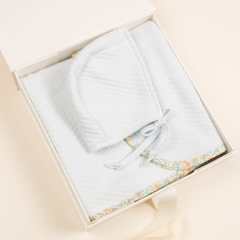 A white baby blanket with a textured pattern is elegantly folded in a Small Gift Box with Bow from the Gift Wrap collection. Accompanied by a matching bonnet with ties, the blanket features floral patterned trim along the edges. This beautiful set, ideal for gift presentation, rests gracefully on a light, neutral surface.