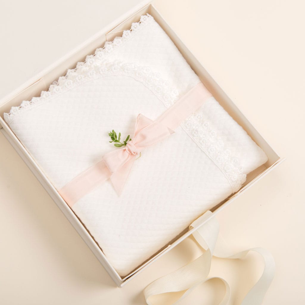The "Gift Wrap | Small Gift Box with Bow" contains a white textured blanket embellished with lace trim, carefully folded inside. It's elegantly wrapped with a pink ribbon tied into a bow and complemented by a small green sprig. Next to the box, there's an elegant cream-colored ribbon from Baby Beau & Belle that enhances the presentation.