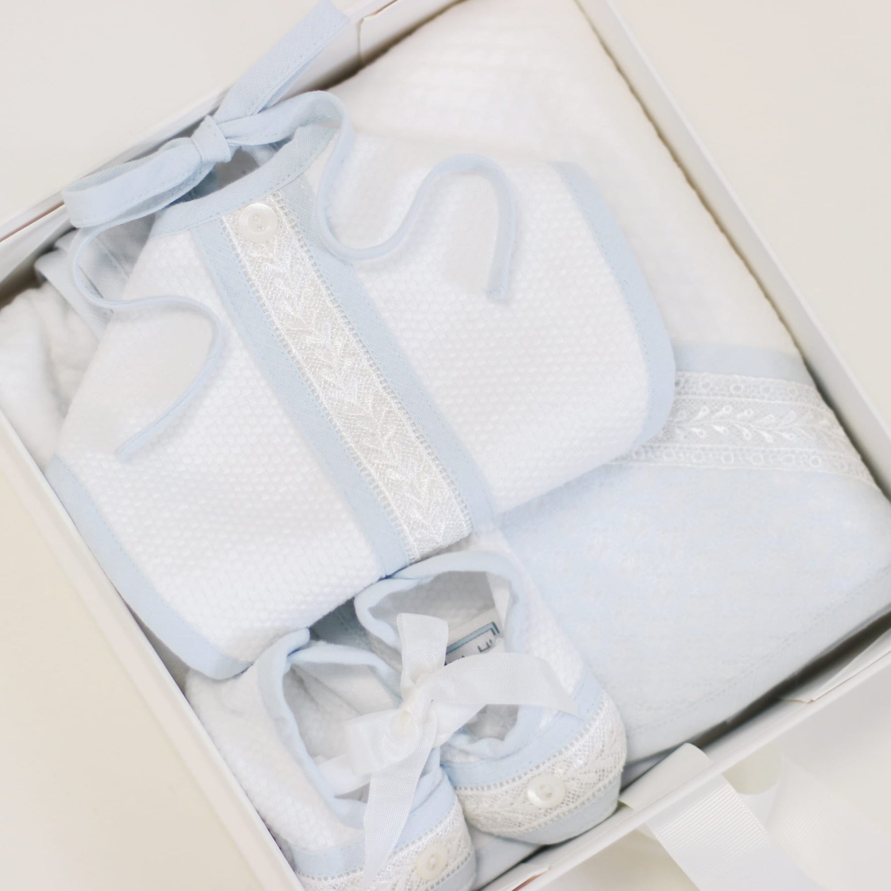 A meticulously arranged Gift Wrap | Small Gift Box with Bow showcases white and light blue baby clothes adorned with lace detailing. This charming set includes a bib, a towel, and a pair of soft shoes, each harmoniously coordinated in color and design for an exquisite gift presentation.