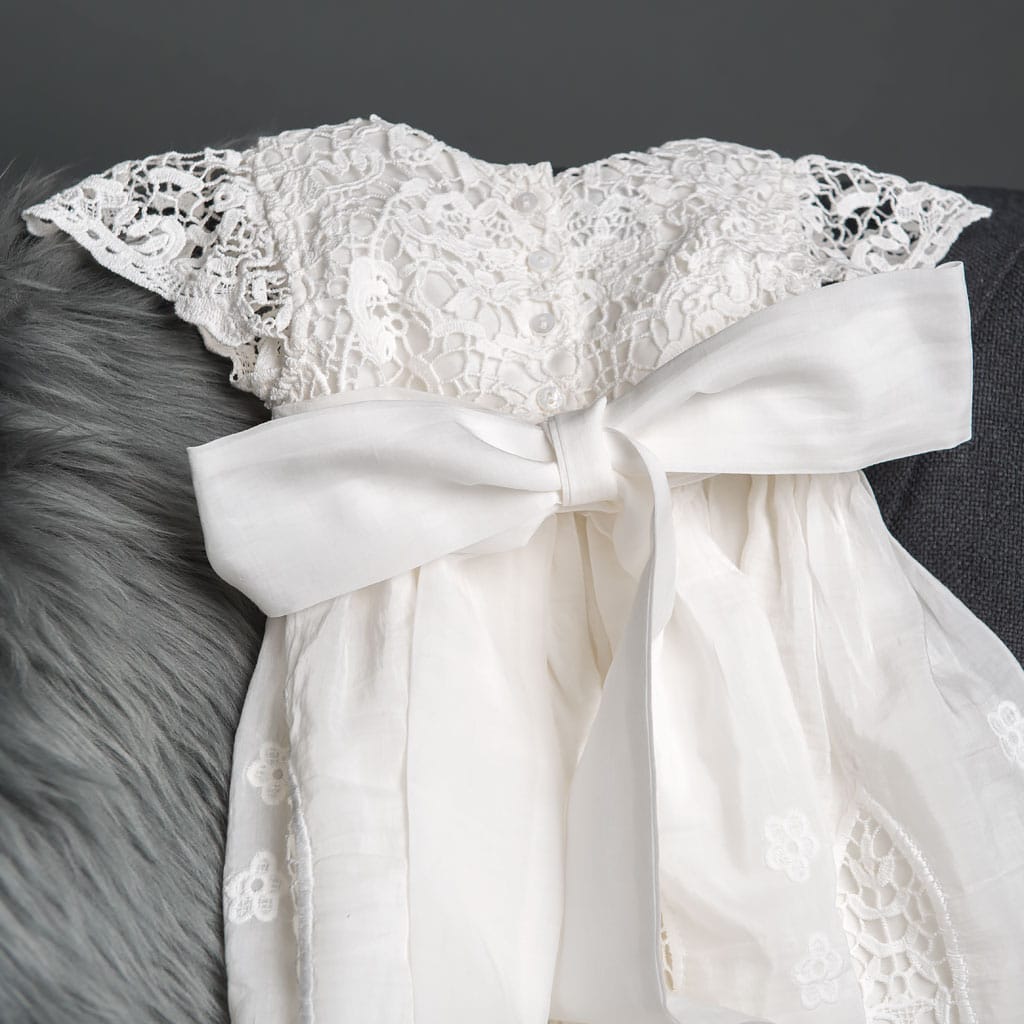 The Grace Christening Gown & Bonnet is showcased on a gray fabric surface, featuring intricate lace detailing on the bodice, with a large white bow adorning the back. The dress has a soft, flowing skirt and is paired with a matching lace Christening Bonnet. The background includes a section of light gray fur fabric.