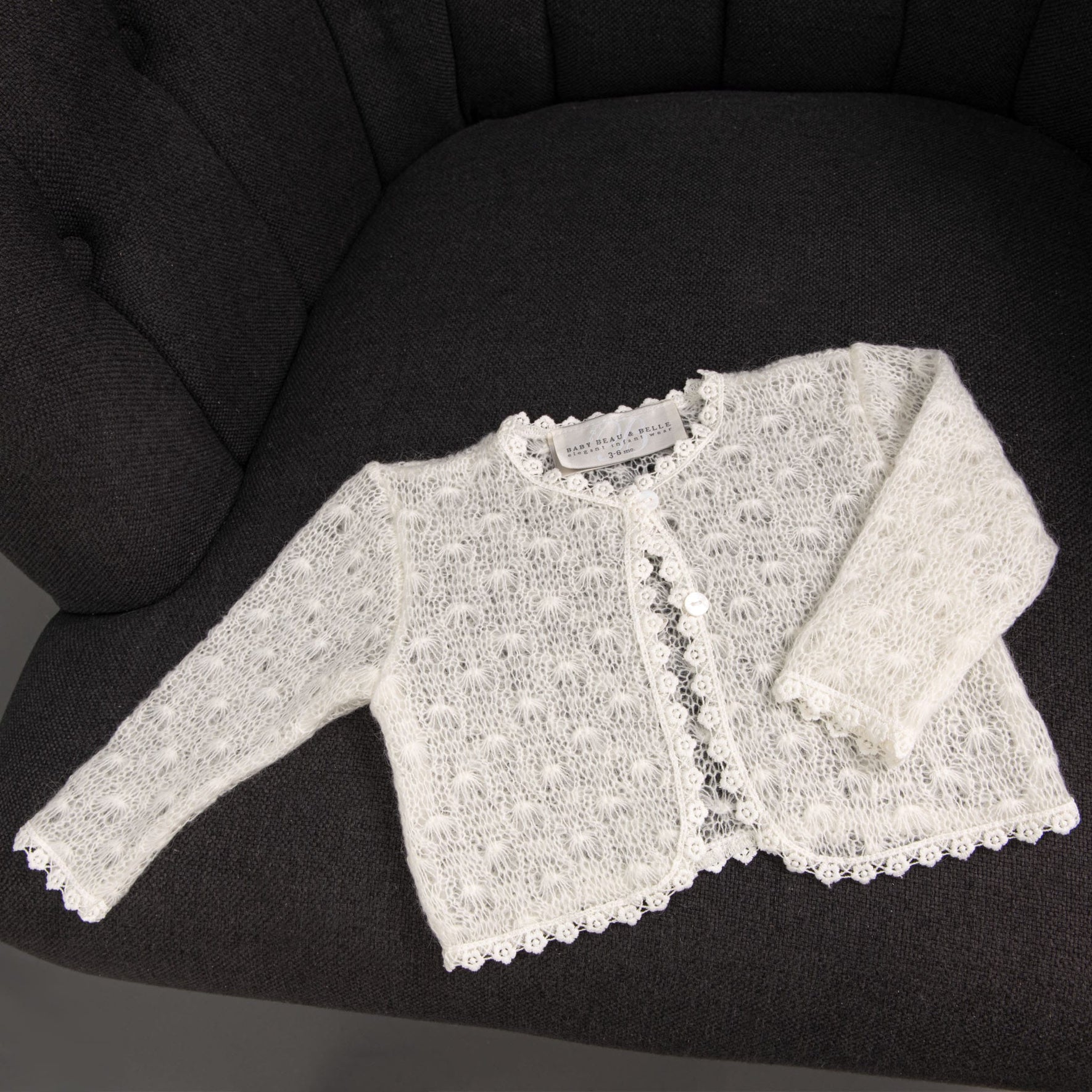 The Grace Christening Knit Sweater, a white knitted baby girls cardigan adorned with cotton floral lace edges, is displayed on a dark upholstered chair. This buttoned-up sweater, ideal for a Christening Day accessory, includes a label inside the collar.