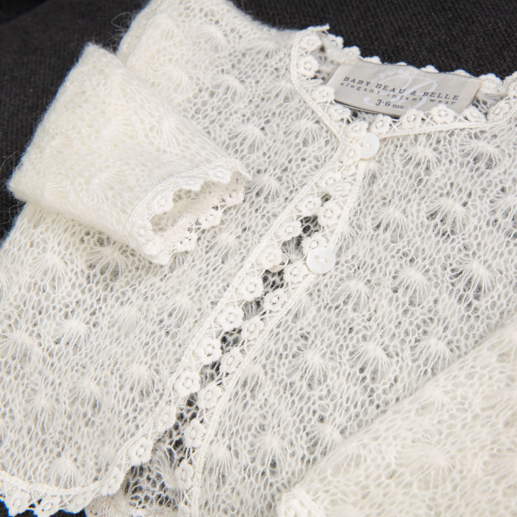 A close-up of the Grace Christening Knit Sweater reveals a white, knitted design with delicate cotton floral lace trim. The fabric appears soft and fluffy with small buttons down the front. Inside the neckline is a label reading "Baby Beau & Belle 3-6 months," making it a perfect Christening Day accessory for baby girls.