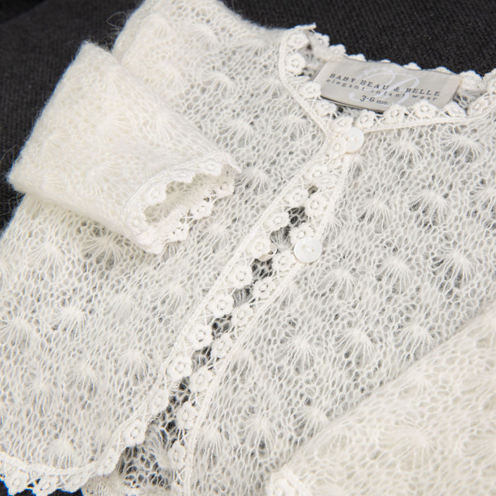A close-up of the Grace Christening Knit Sweater reveals a white, knitted design with delicate cotton floral lace trim. The fabric appears soft and fluffy with small buttons down the front. Inside the neckline is a label reading "Baby Beau & Belle 3-6 months," making it a perfect Christening Day accessory for baby girls.