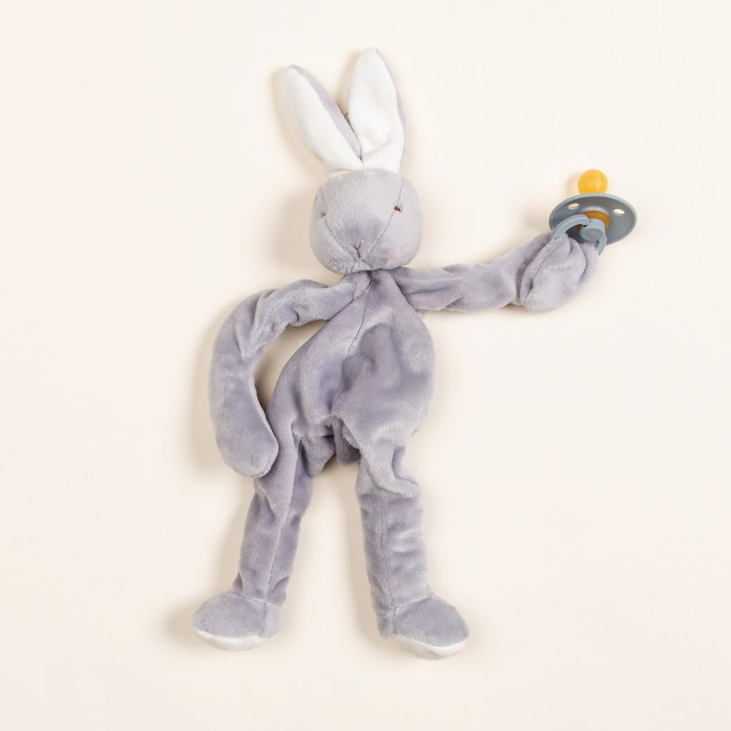 The Grey Silly Bunny Buddy, a plush rabbit with extended limbs and white ears, gently holds a pacifier featuring a yellow top and gray base against a simple backdrop. This makes it an ideal gift for baby showers.