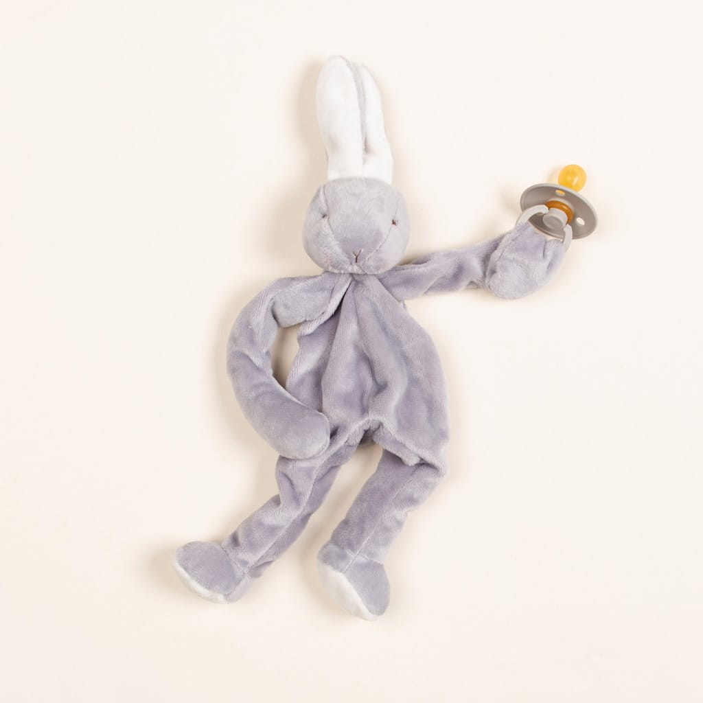The Grey Silly Bunny Buddy, a plush grey rabbit toy with long ears, holds a pacifier featuring a grey ring and orange nipple in its left hand. Ideal for gifting at baby showers, this charming bunny is displayed against a simple light background.